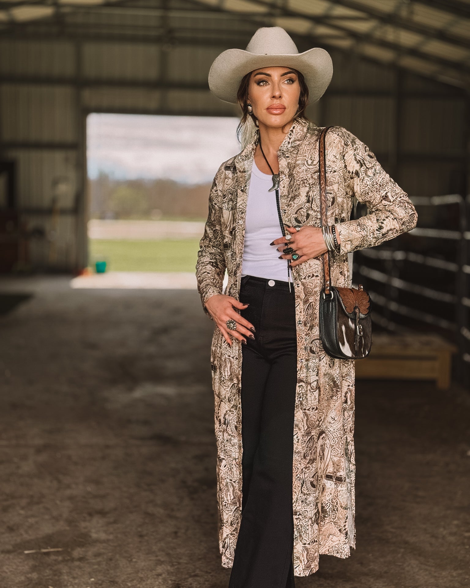 Woman in a western boho outfit with a cowboy hat, long patterned duster, and black flared pants standing in a barn.