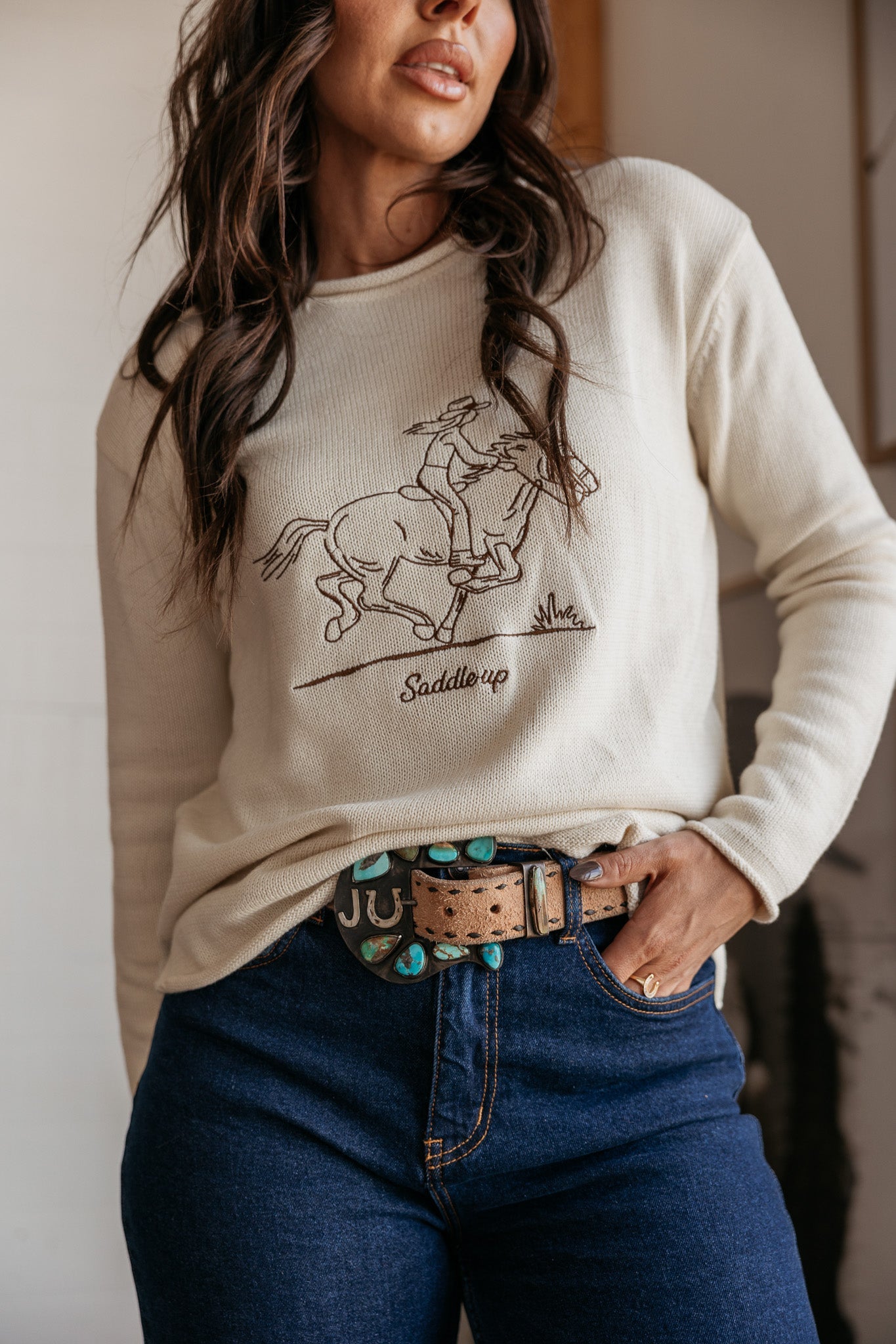 Woman wearing a cream western graphic sweatshirt with 'Saddle up' text, turquoise concho belt, and blue jeans