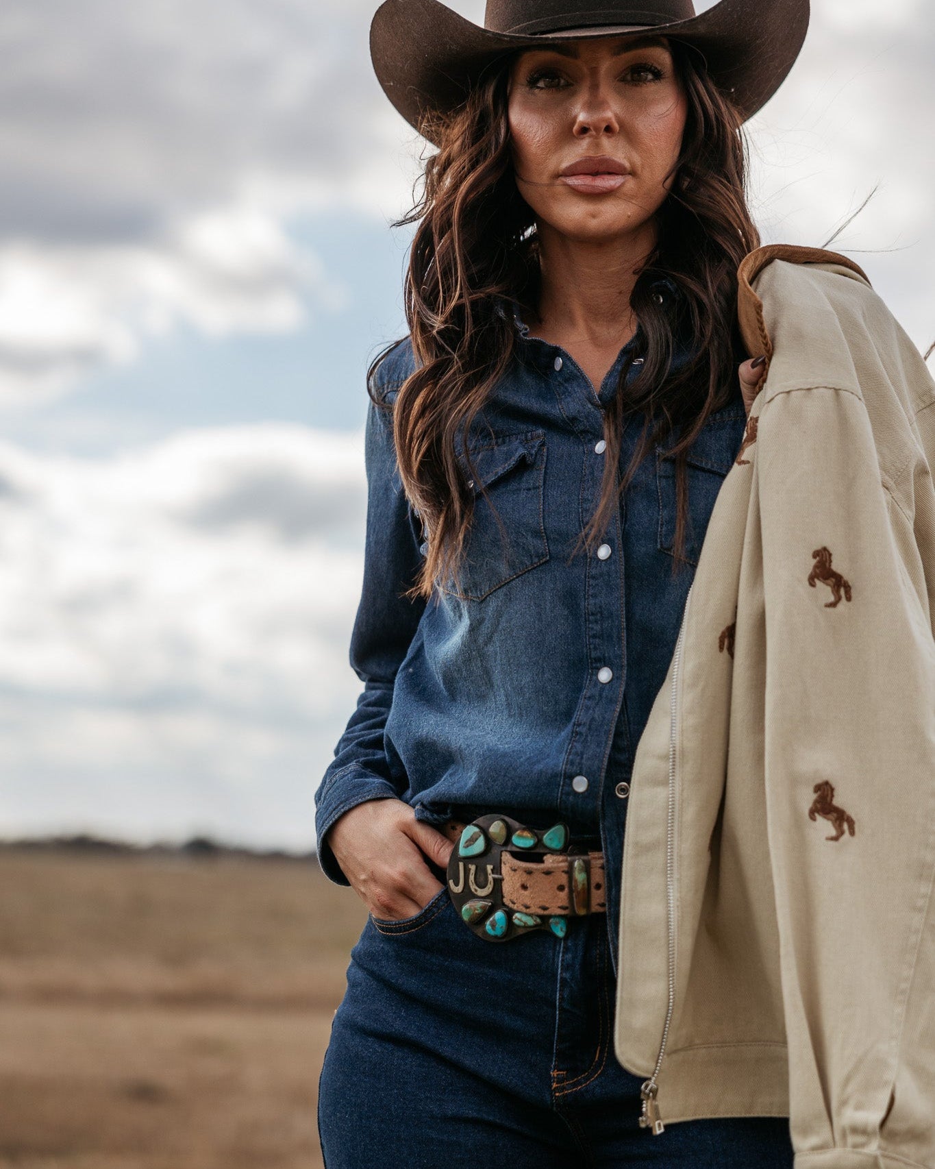 Woman in cowgirl hat and denim shirt wearing turquoise western belt, holding tan jacket, outdoors