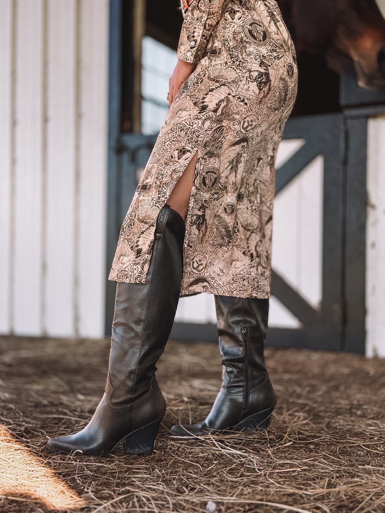 Woman in western print dress and black cowgirl boots at barn with hay floor