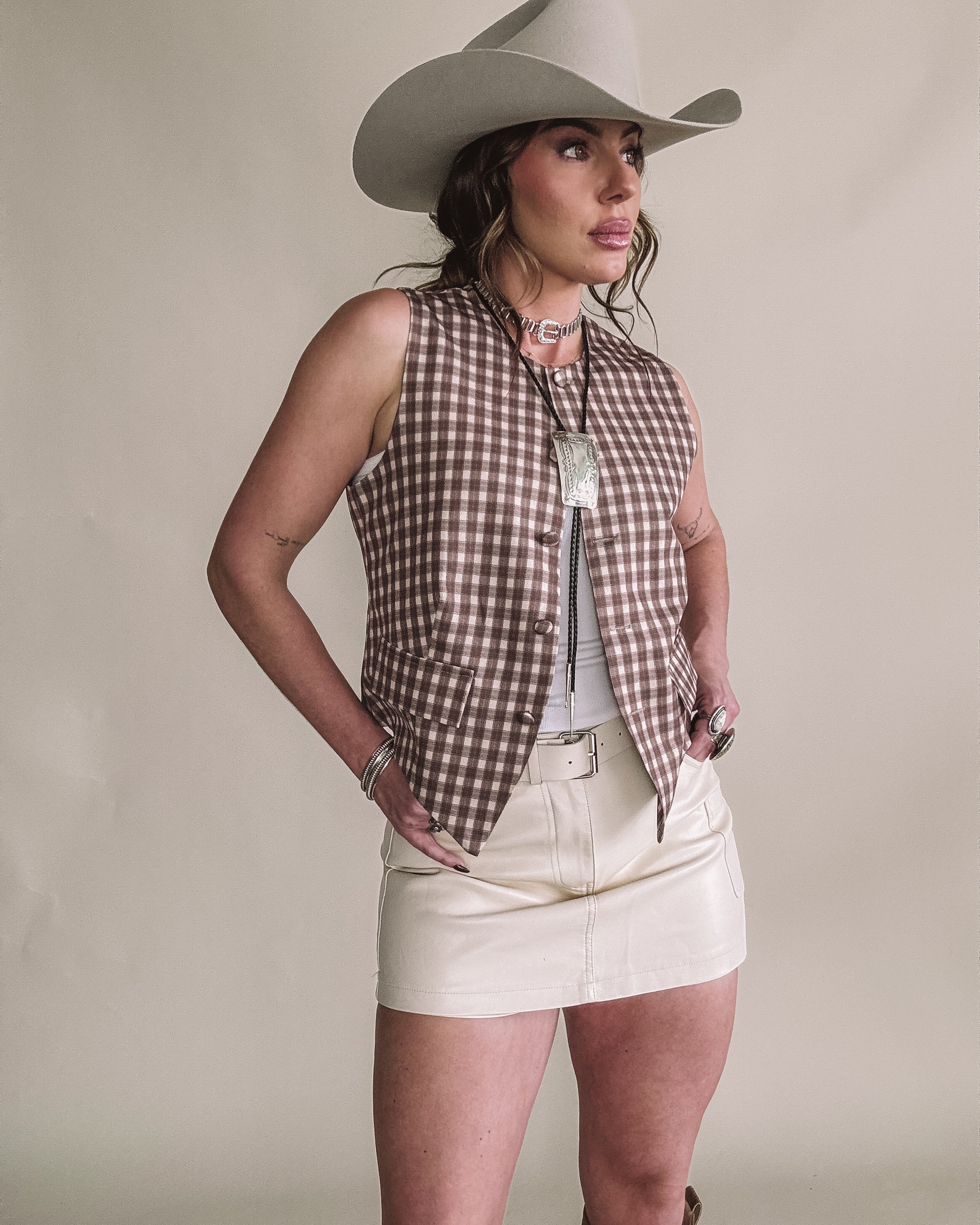 Woman in western outfit with beige cowboy hat, plaid vest, cream mini skirt, and statement necklace.