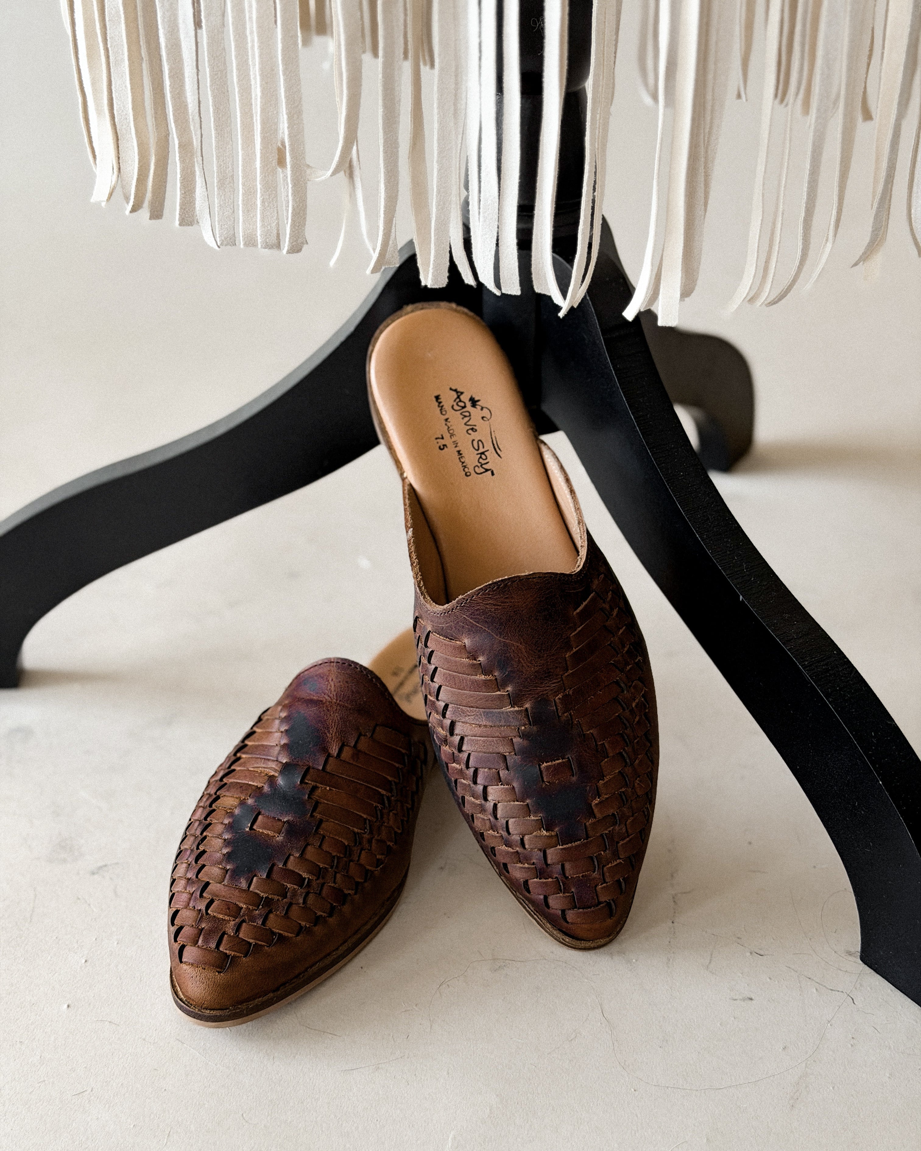 Brown woven leather western mules on floor with white fringe decor in background