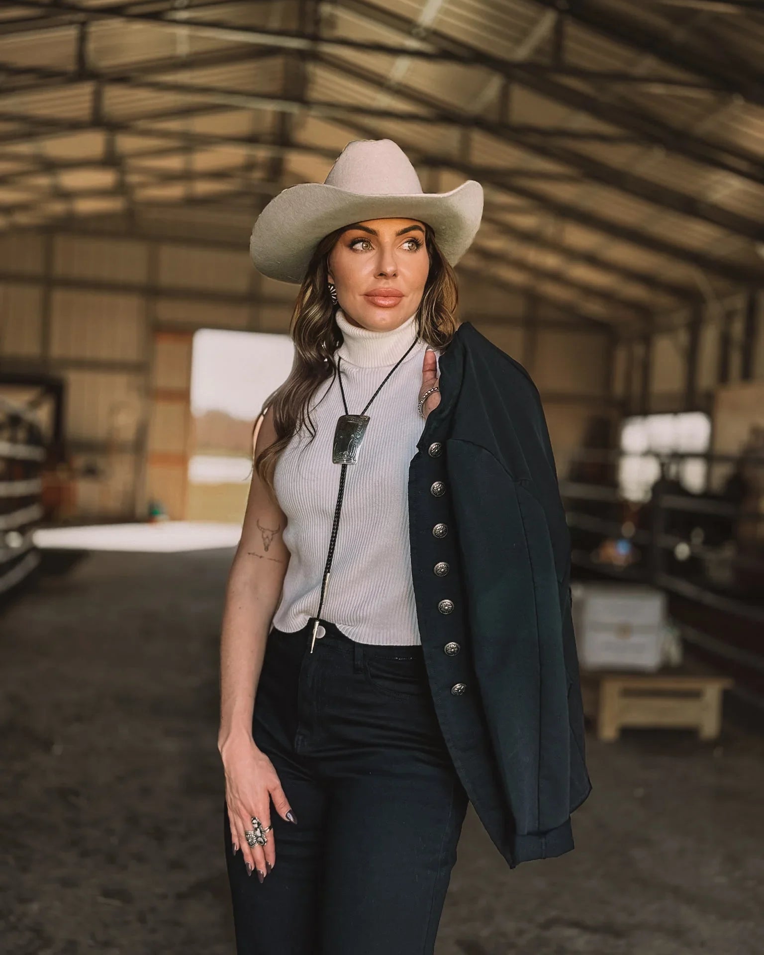 Woman in cowgirl hat, western necklace, white sleeveless top, black pants and jacket, standing in barn