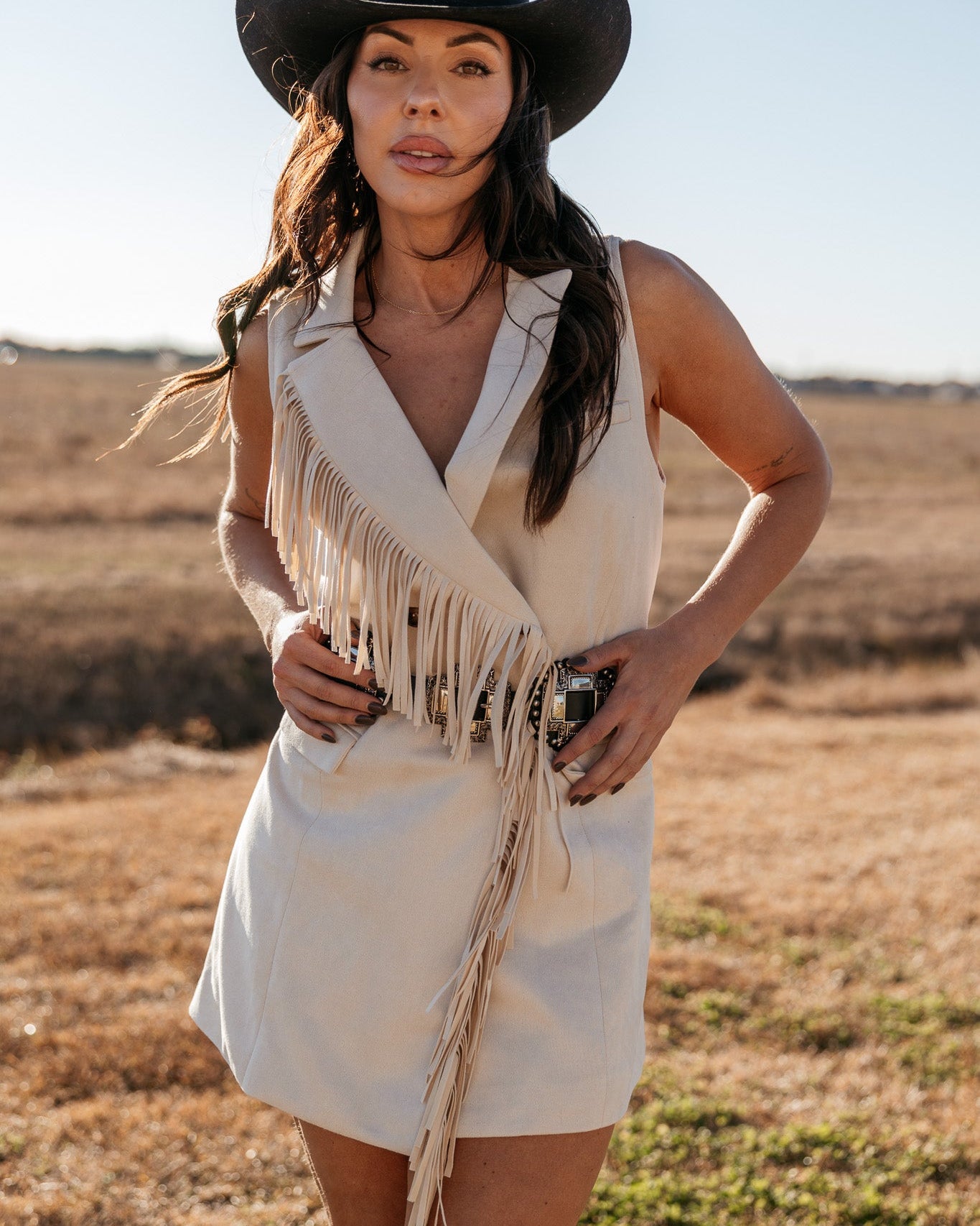 Woman in western fringe dress and cowboy hat posing outdoors in a boho country outfit.