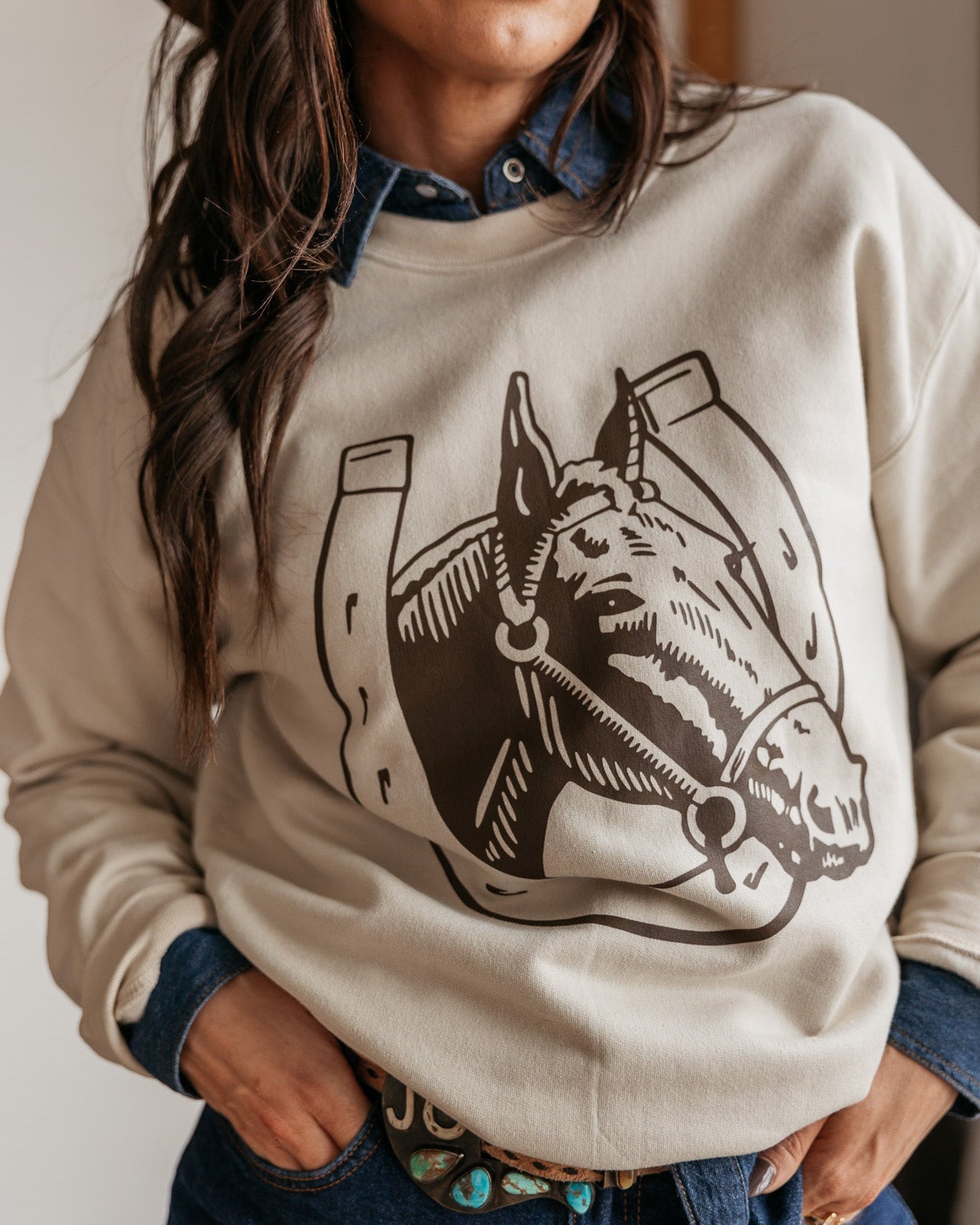 Woman in western graphic sweatshirt with horse and horseshoe, denim shirt, and turquoise belt