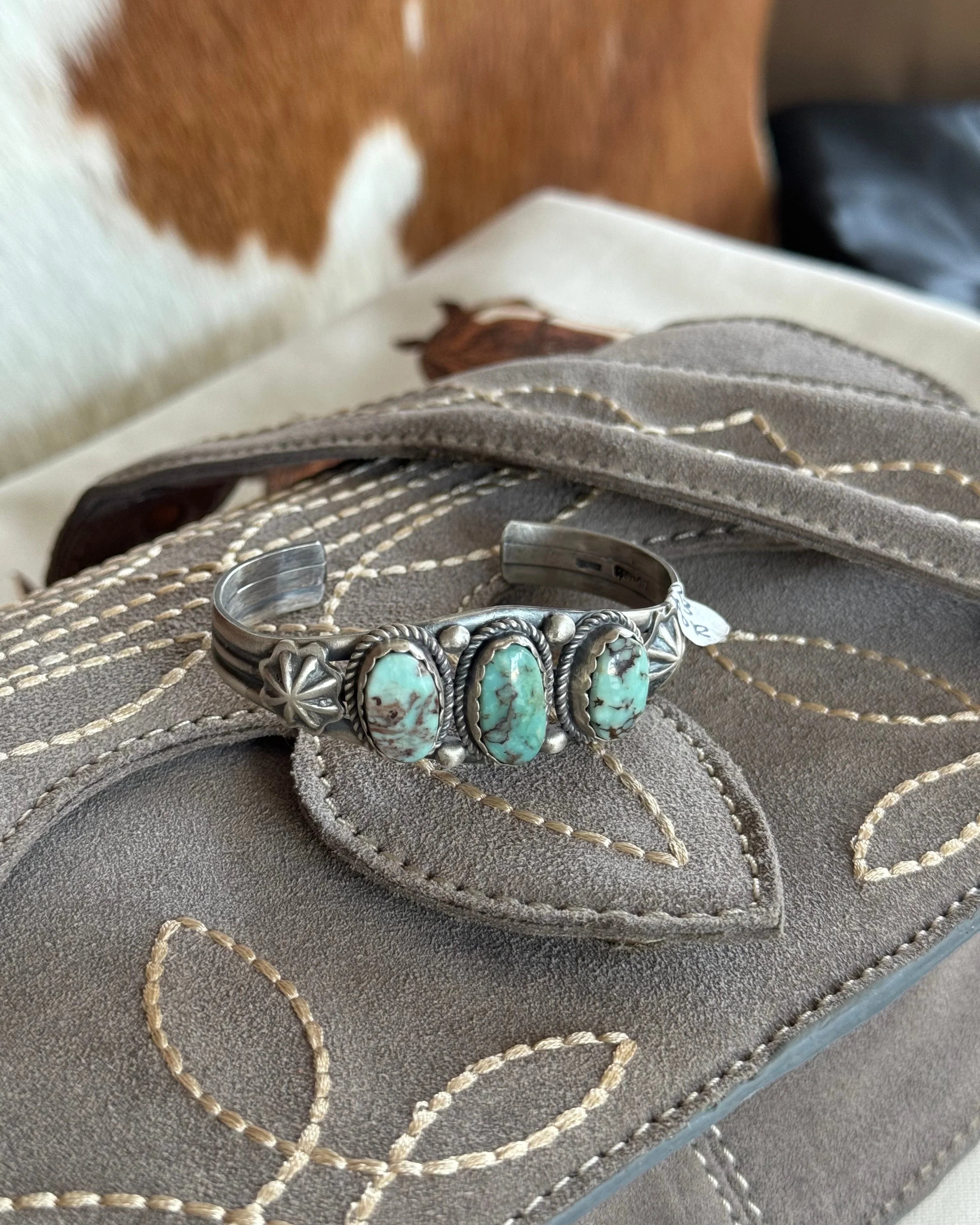 Western turquoise cuff bracelet on embroidered suede clutch, cowhide background.