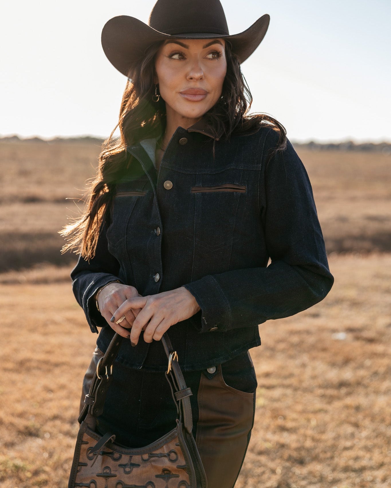 Woman in western denim jacket and black cowboy hat holding a patterned saddle bag outdoors