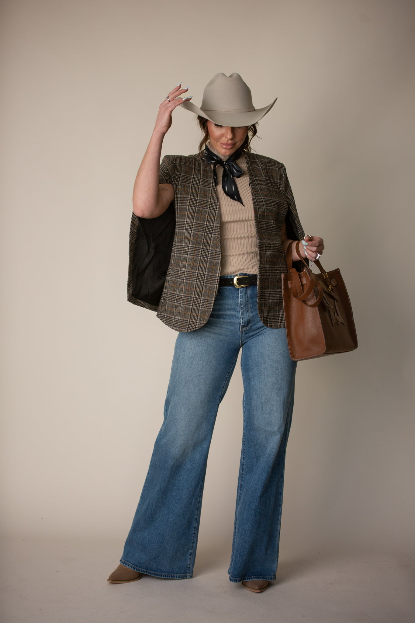Woman in western boho outfit with cowboy hat, plaid blazer, flared jeans, brown handbag