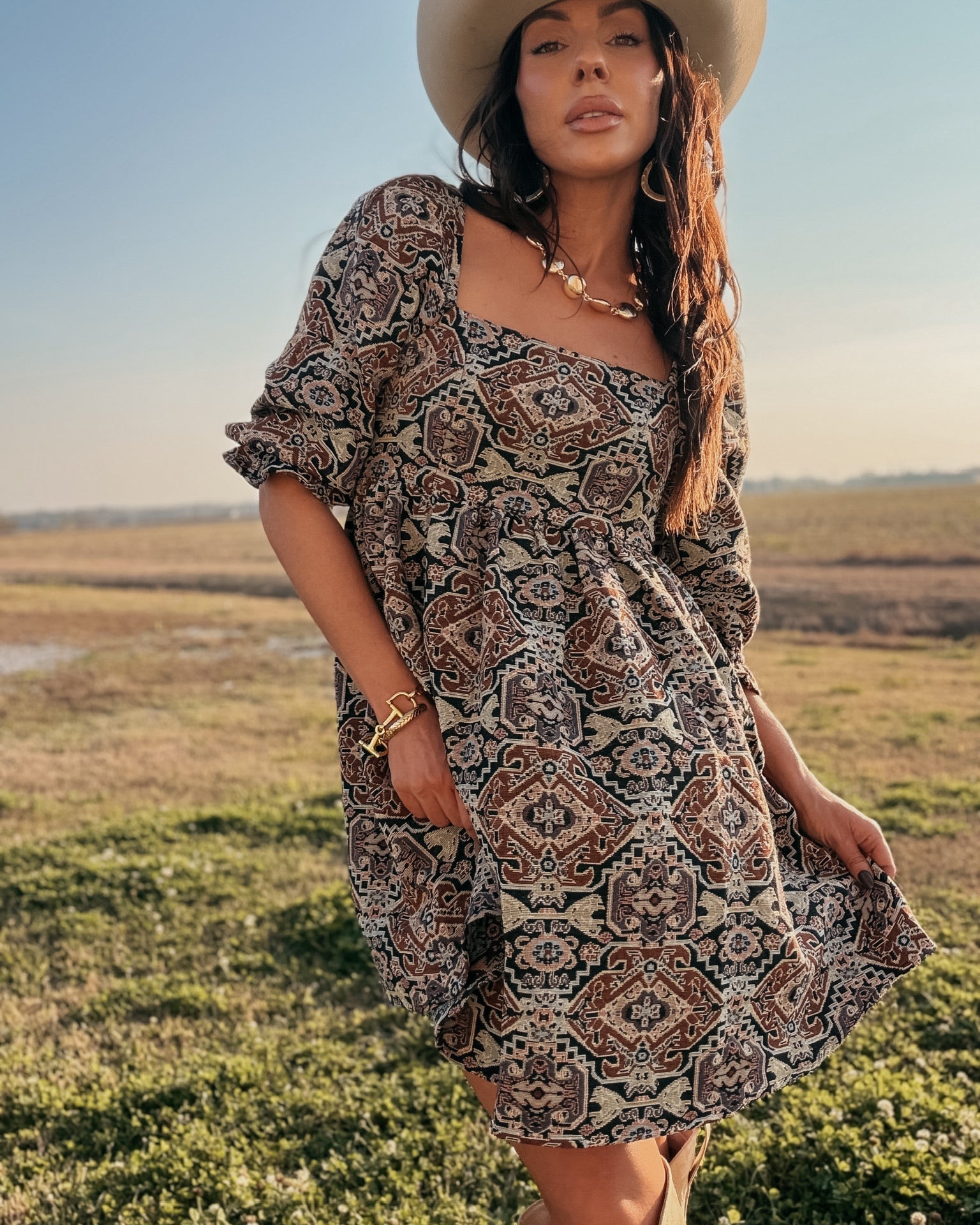 Woman in boho western print dress, cowboy hat, and boots standing outdoors in a field