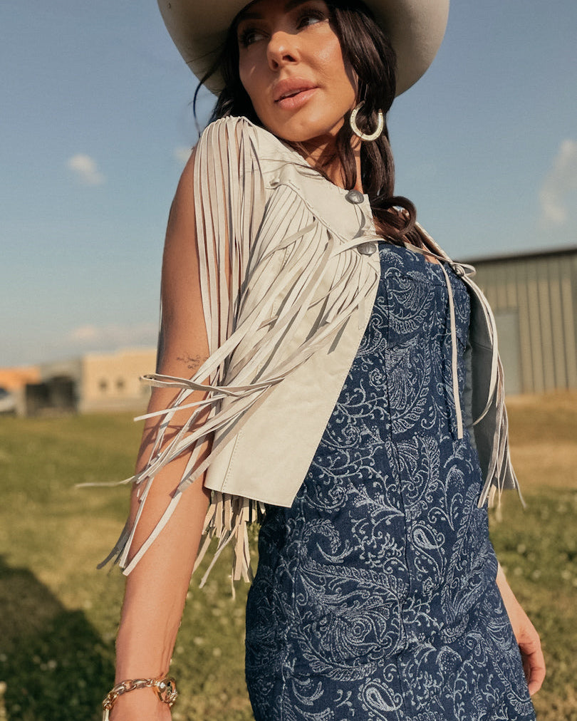 Woman in white fringed vest, blue paisley western dress, and cowgirl hat outdoors