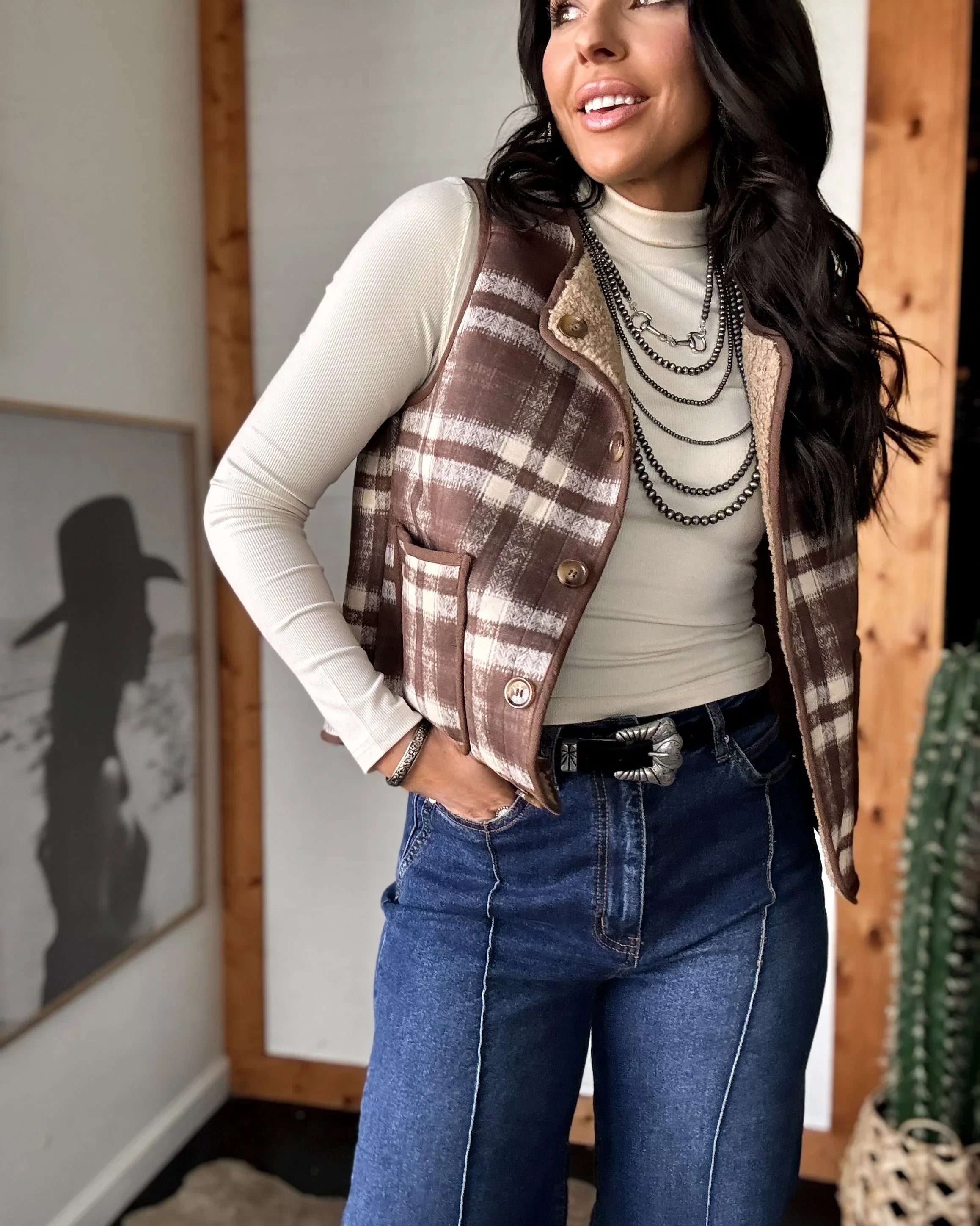 Woman wearing a plaid vest, white turtleneck, and blue jeans indoors.