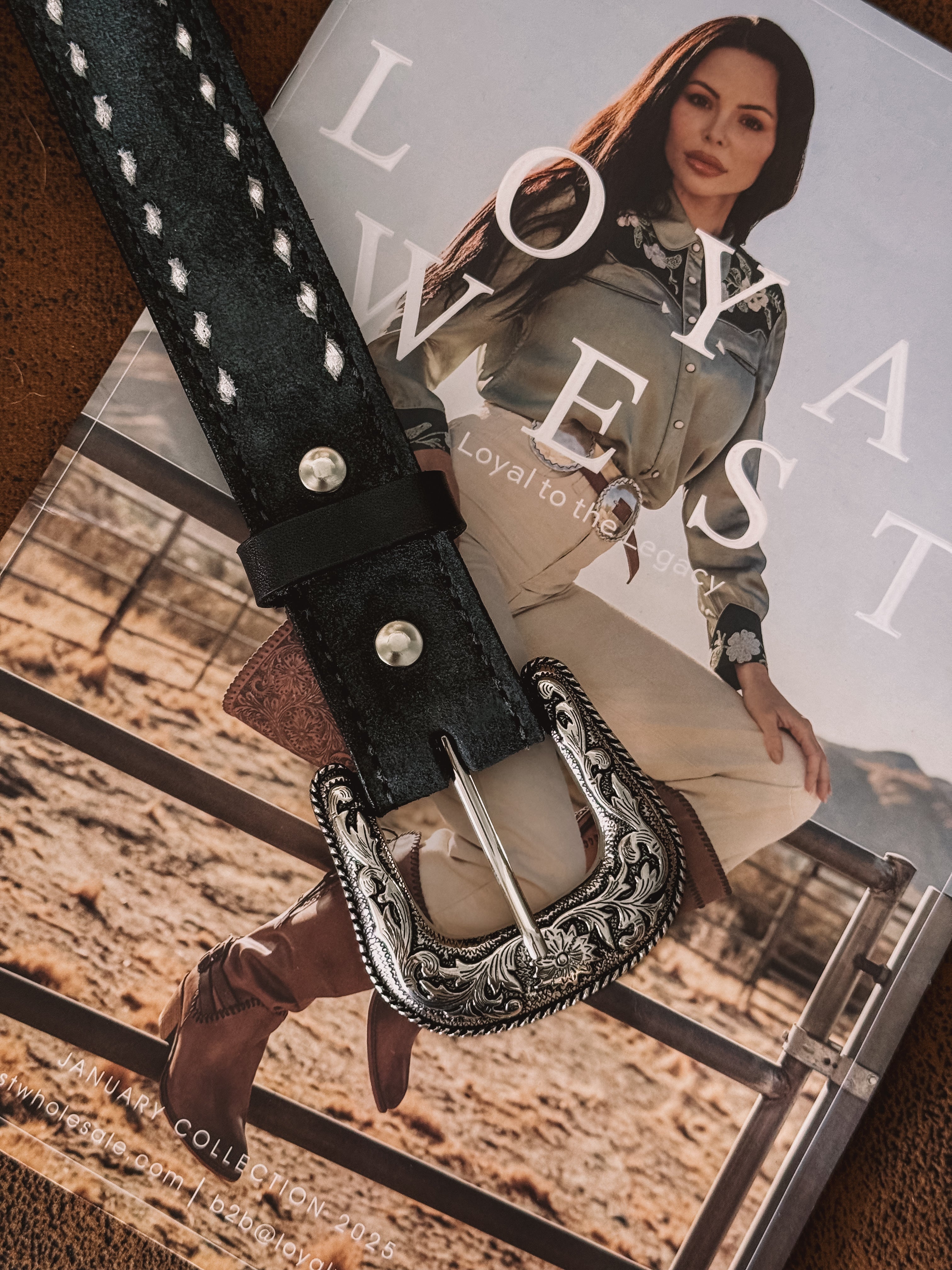 Western engraved silver belt buckle on black leather belt over magazine with cowgirl in rodeo outfit