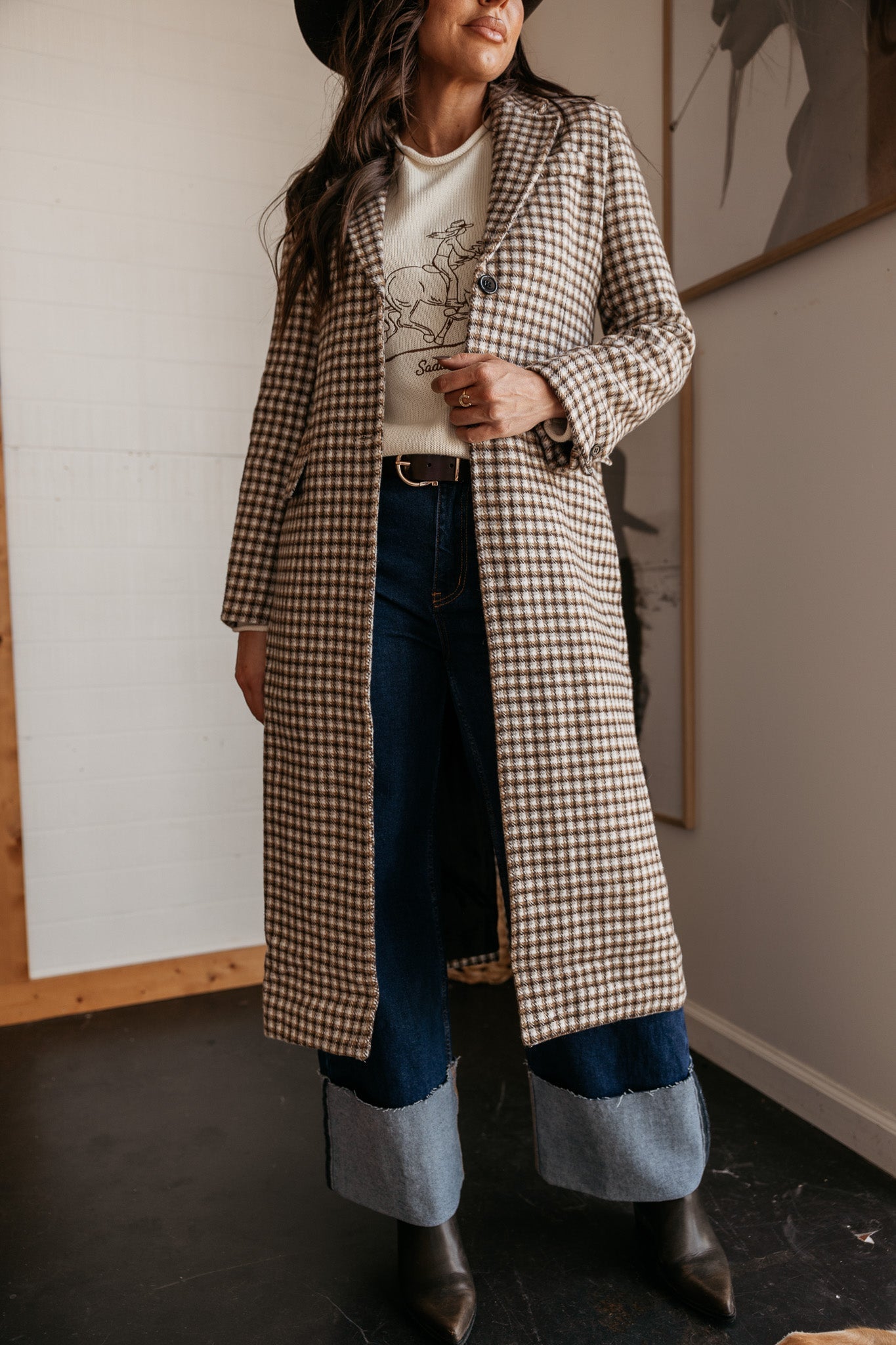 Woman in western boho plaid long coat, graphic sweater, two-tone jeans and cowboy hat