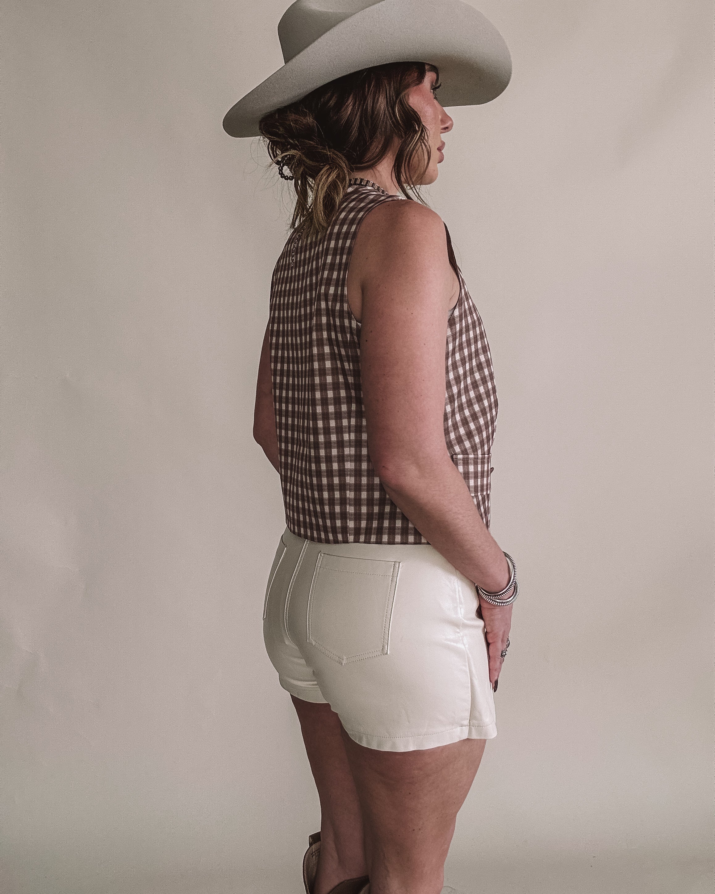 Woman in a western hat, checkered sleeveless top, white shorts, and cowgirl boots