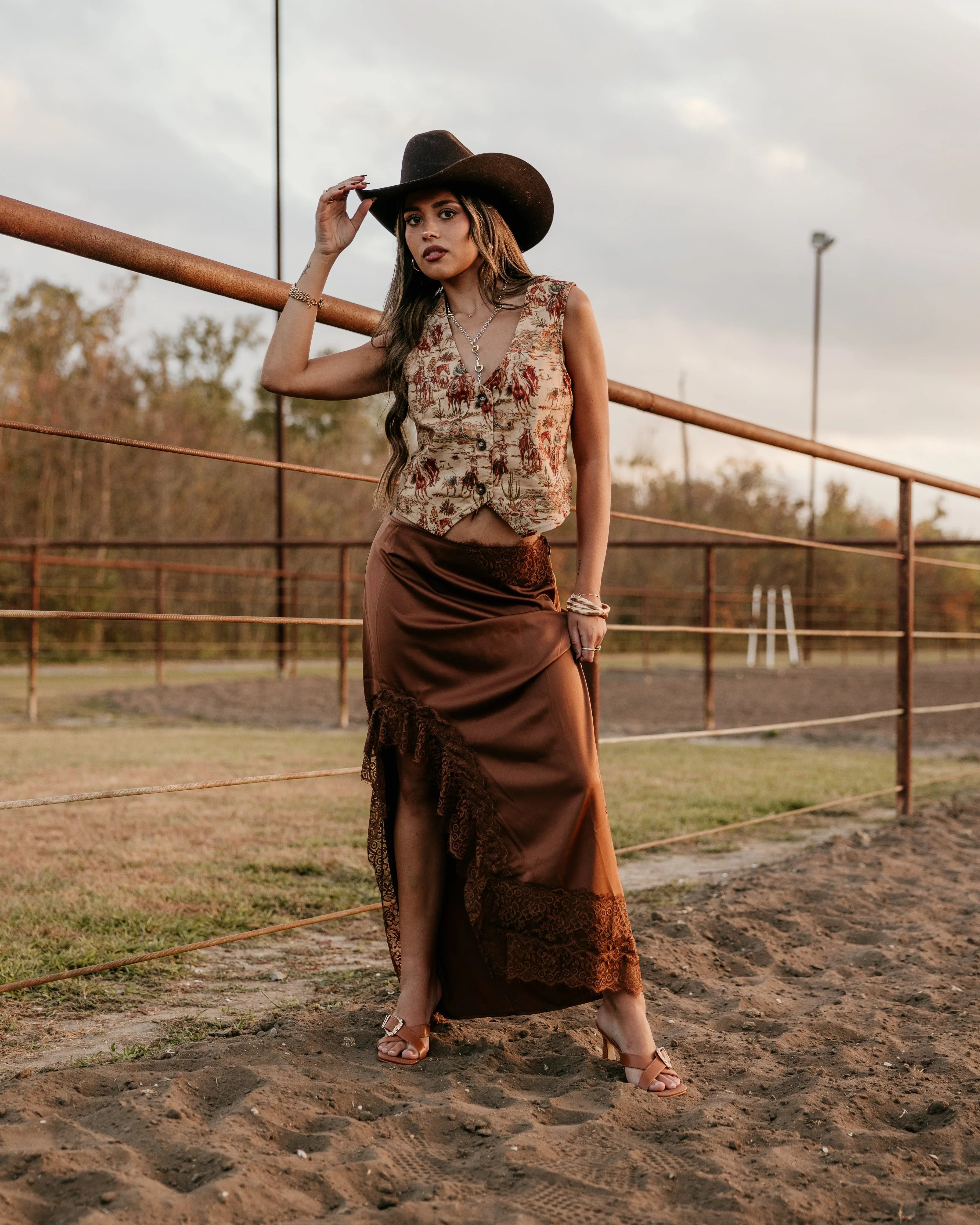 Woman in a sleeveless top and long skirt standing on a dirt path with a fence and trees in the background.