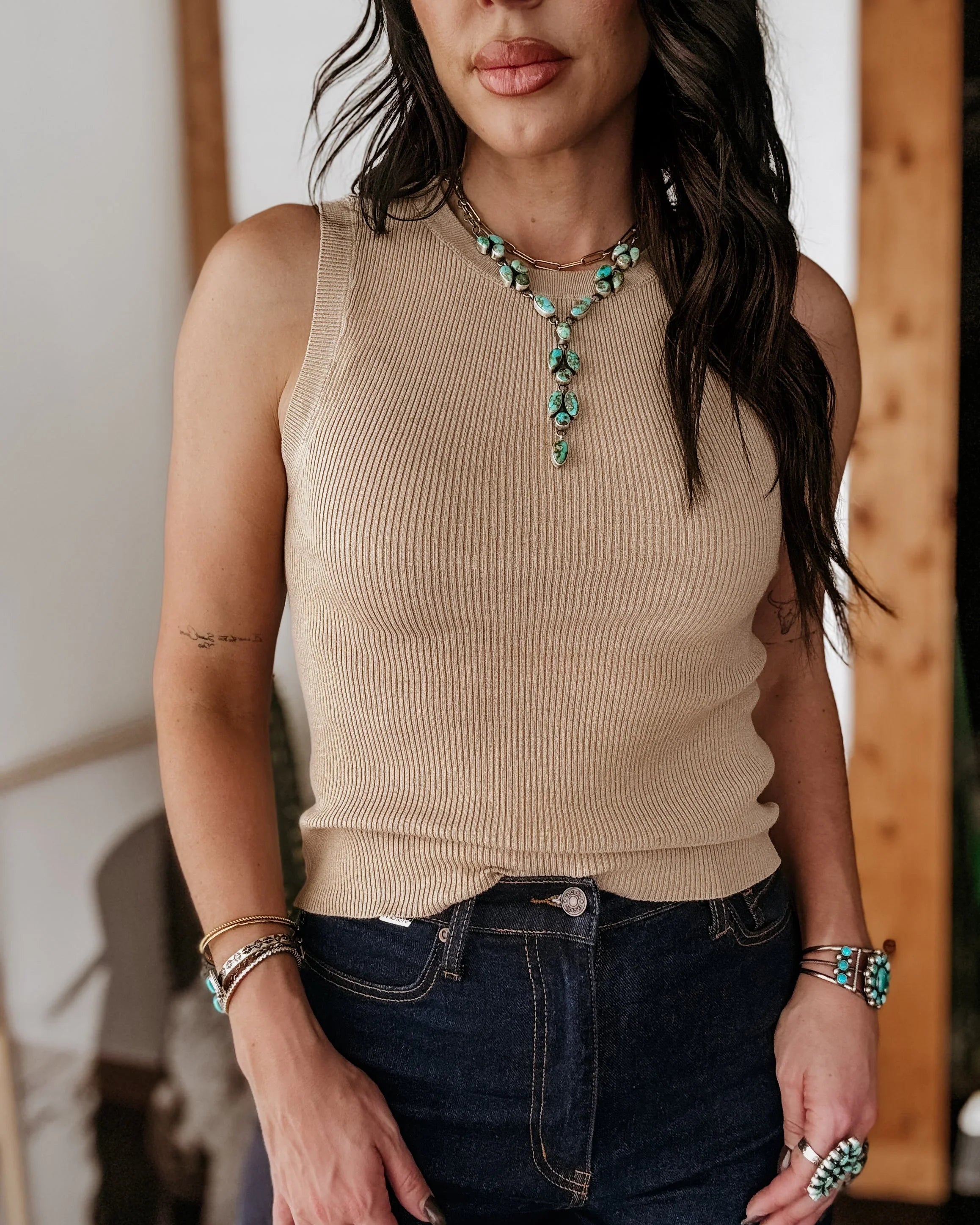 Woman wearing a beige sleeveless top and dark jeans with jewelry.