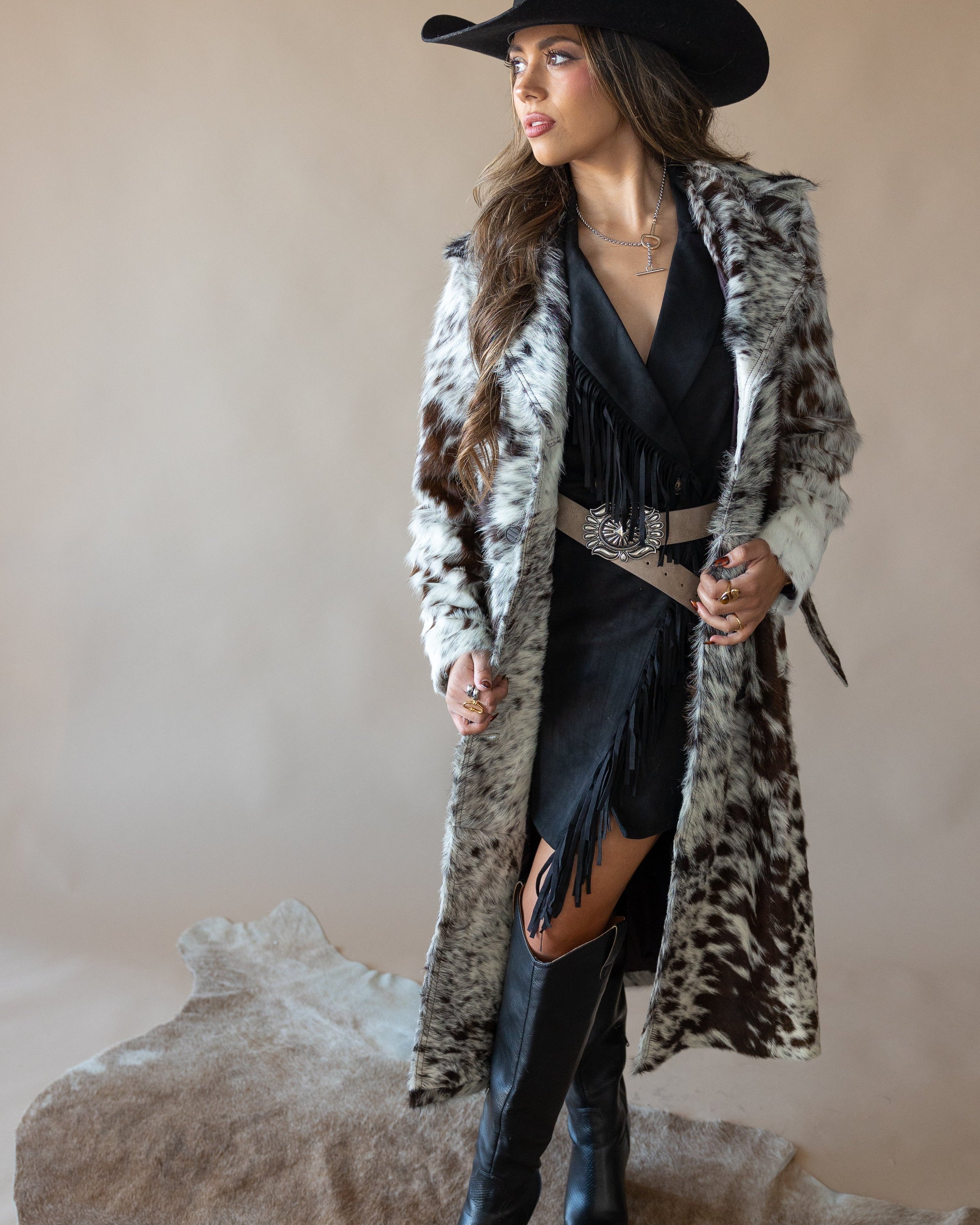 Woman in cow print faux fur coat, black fringe dress, cowboy hat, and western boots