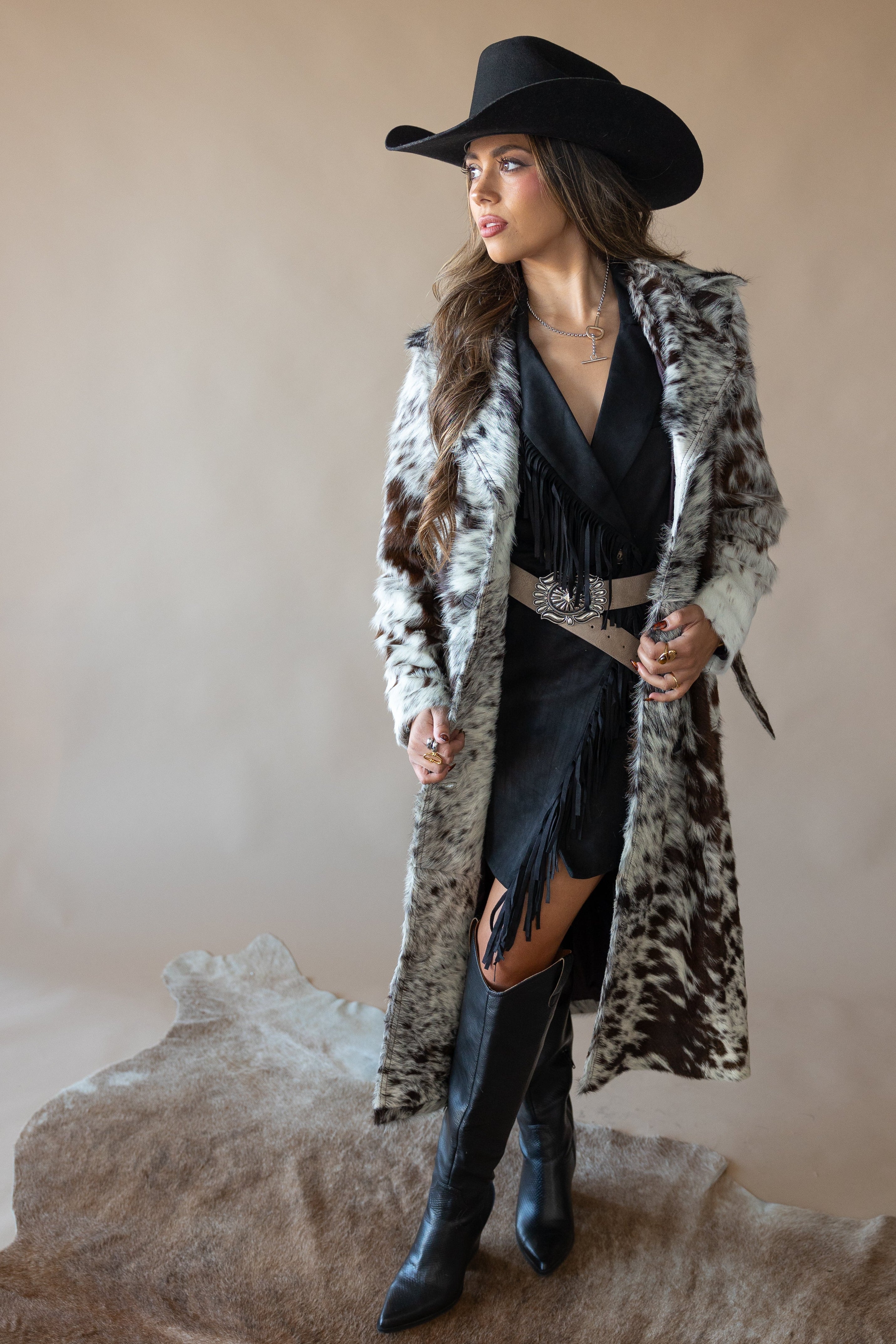Woman in cow print faux fur coat, black fringe dress, cowboy hat, and western boots