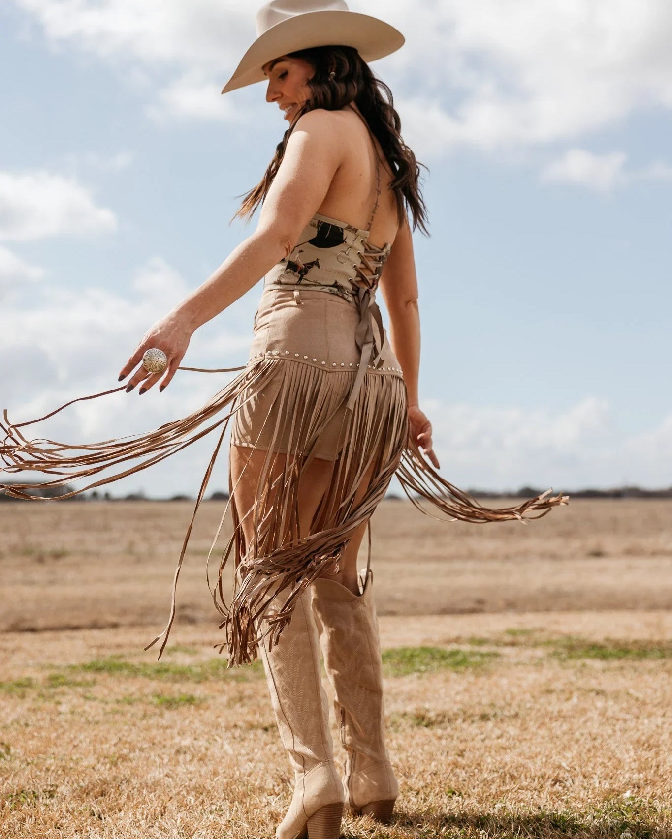 Woman in western boho outfit with fringe skirt, cowboy hat, and boots standing outdoors