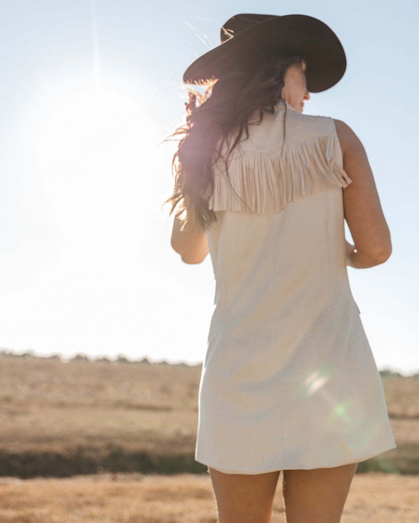 Woman in western fringe dress, cowboy hat, and boots standing outdoors in sunlight