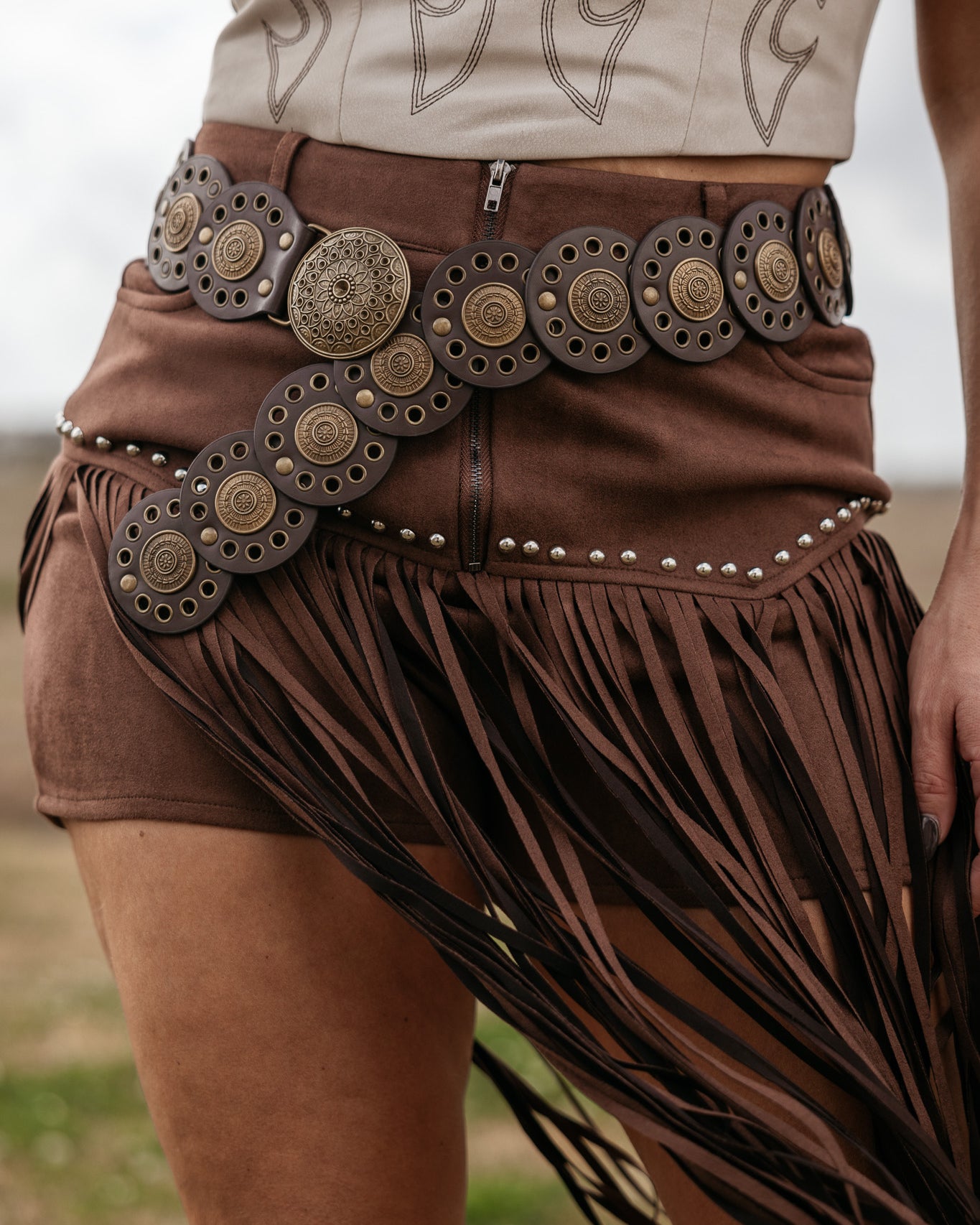 Brown fringe skirt with decorative belt worn by a person, against a blurred natural background.