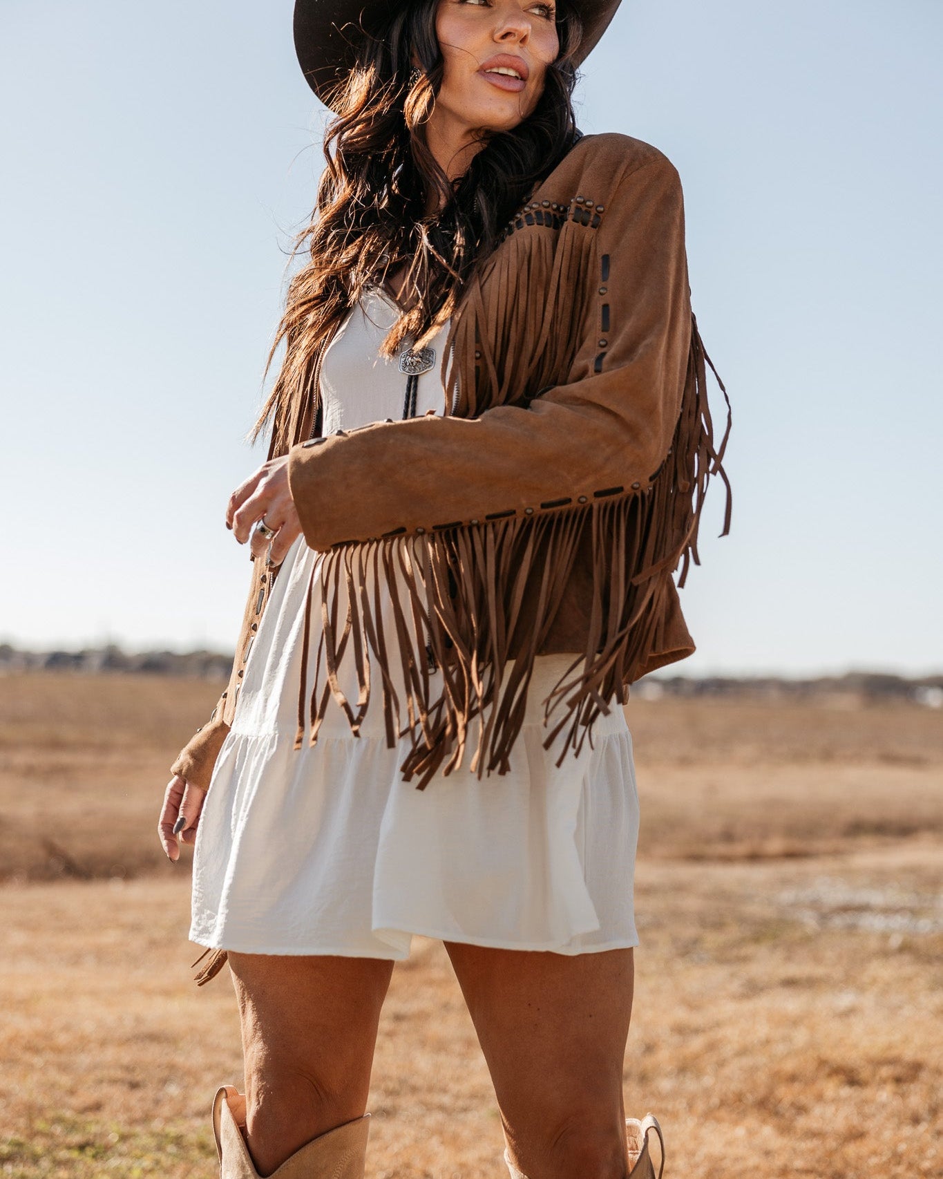 Woman in Western boho outfit with fringe jacket, white dress, cowboy hat and boots outdoors