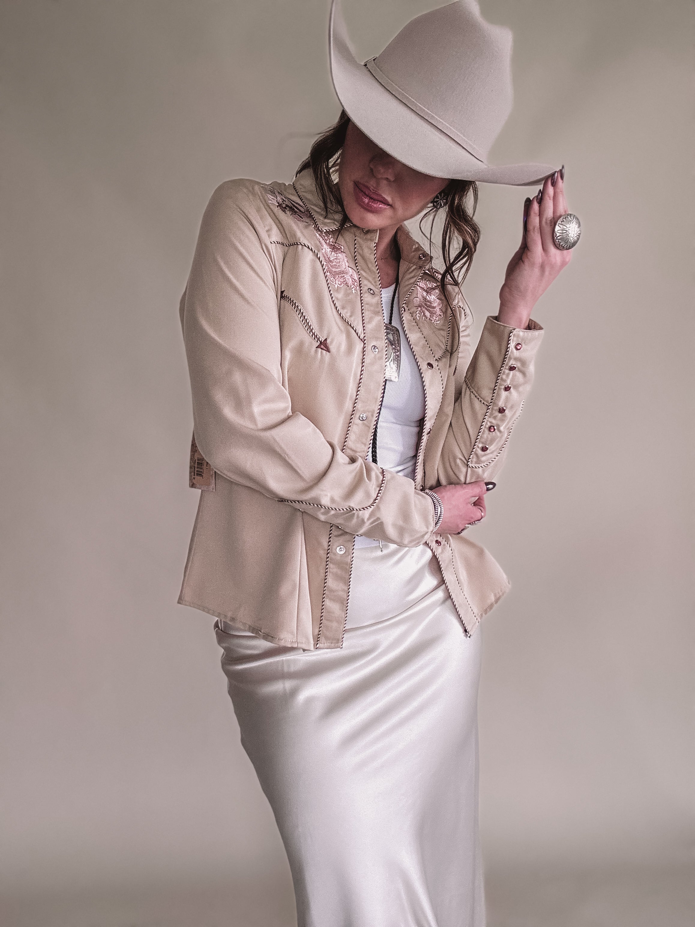 Woman in beige embroidered western shirt, satin skirt, and cowboy hat, showcasing boho cowgirl style.