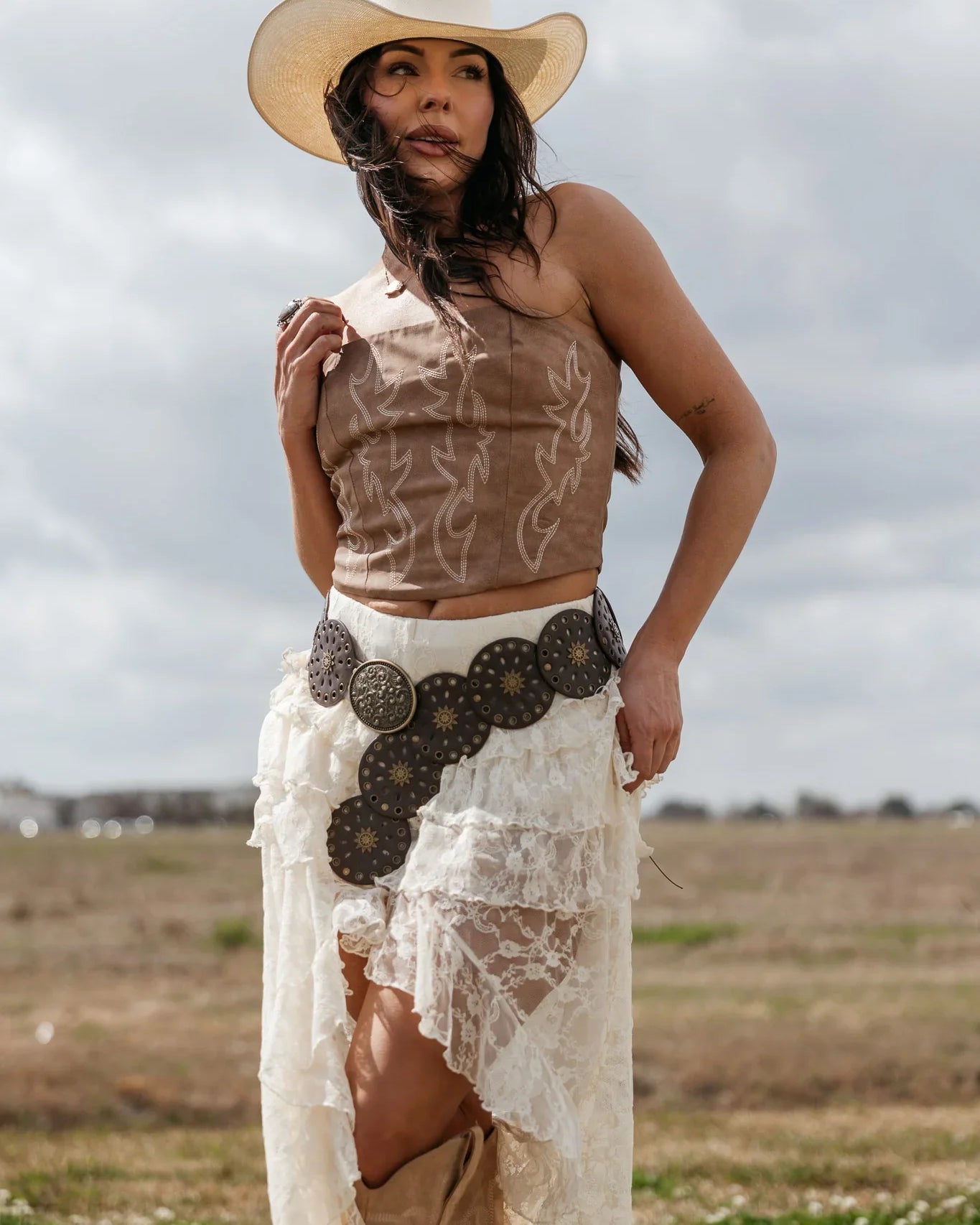 Woman in western boho outfit with cowboy hat, brown corset, lace skirt, and concho belt outdoors