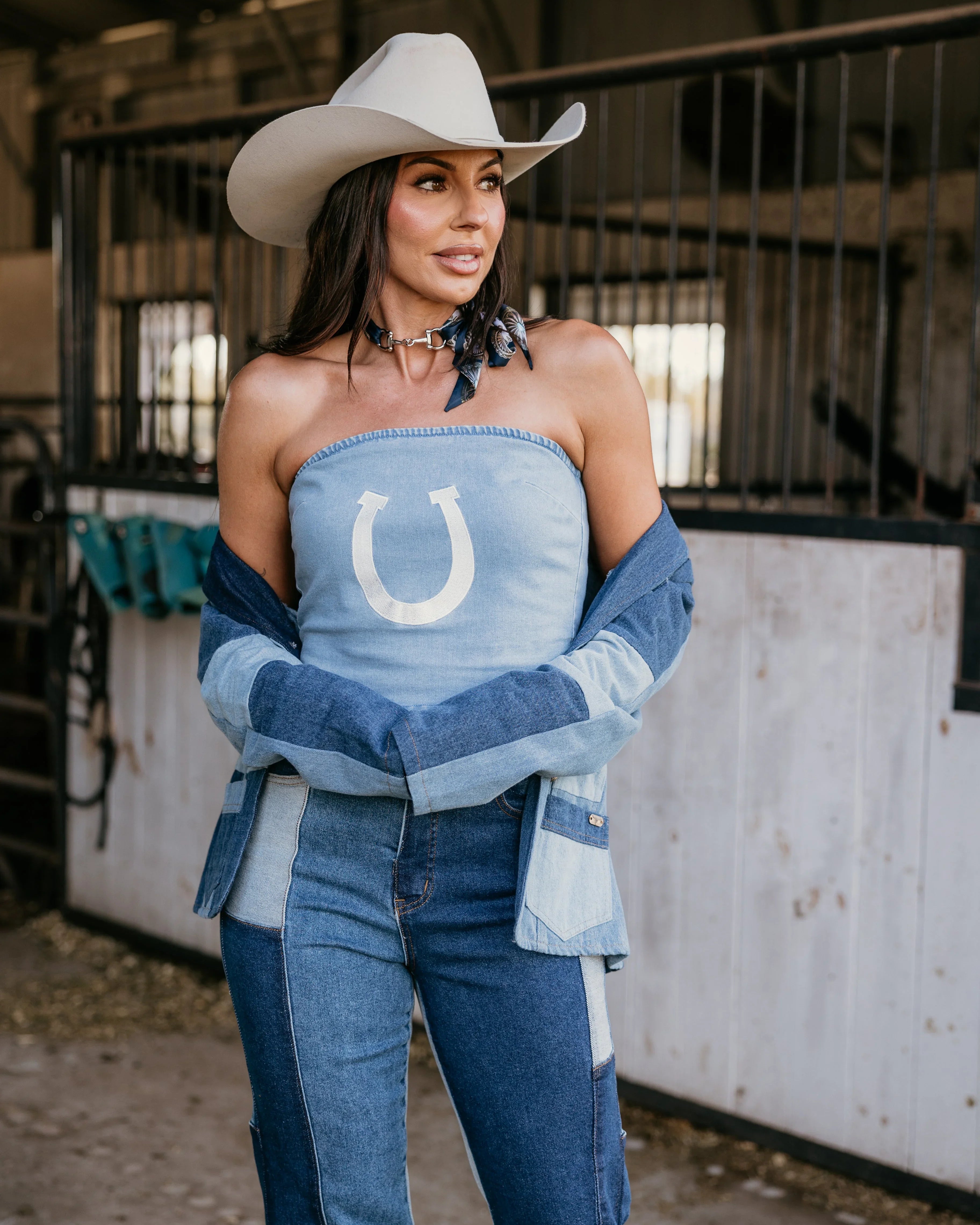 Woman in western boho denim outfit and cowboy hat standing in barn, patchwork jeans, horseshoe top.