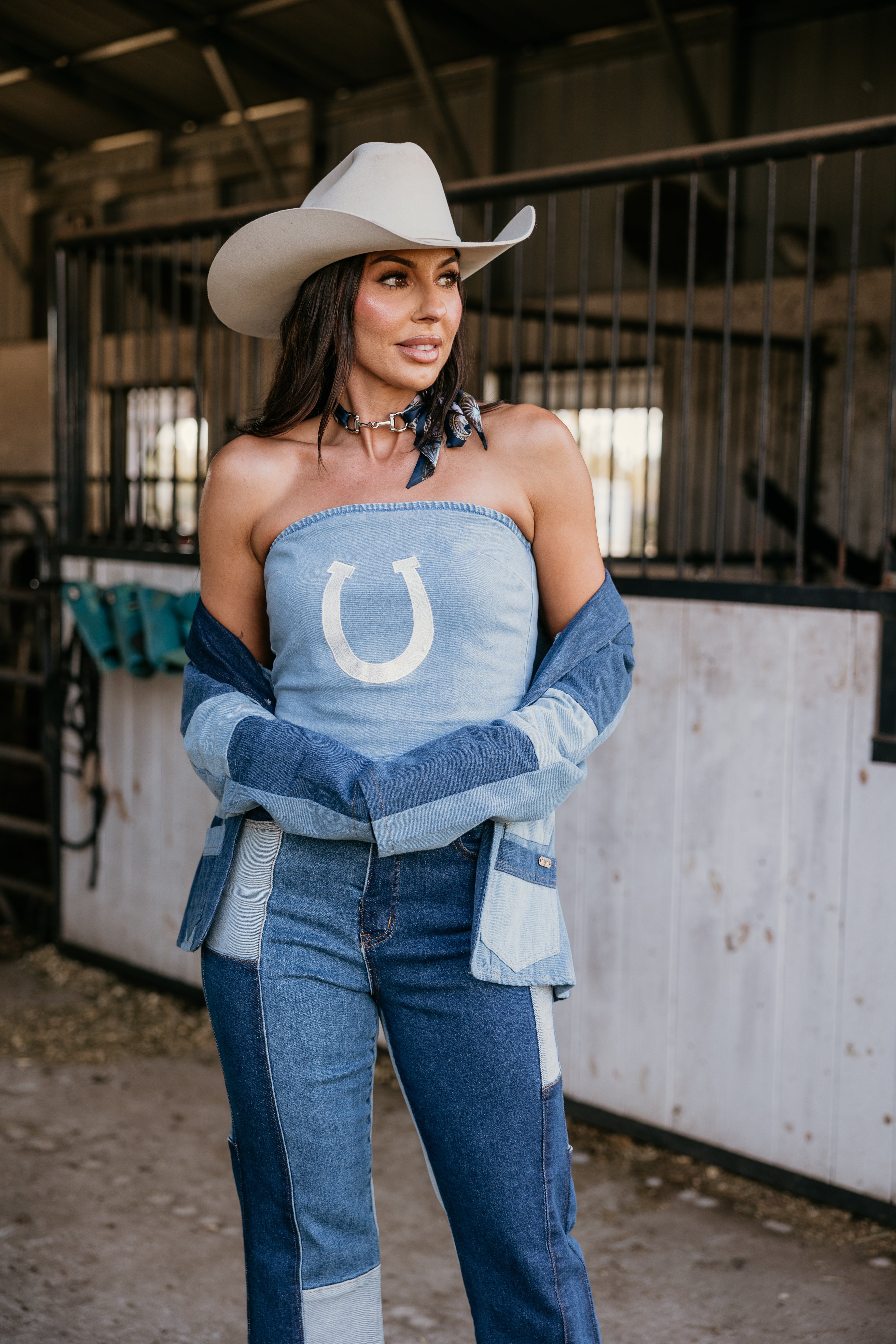 Woman in western boho denim outfit and cowboy hat standing in barn, patchwork jeans, horseshoe top.