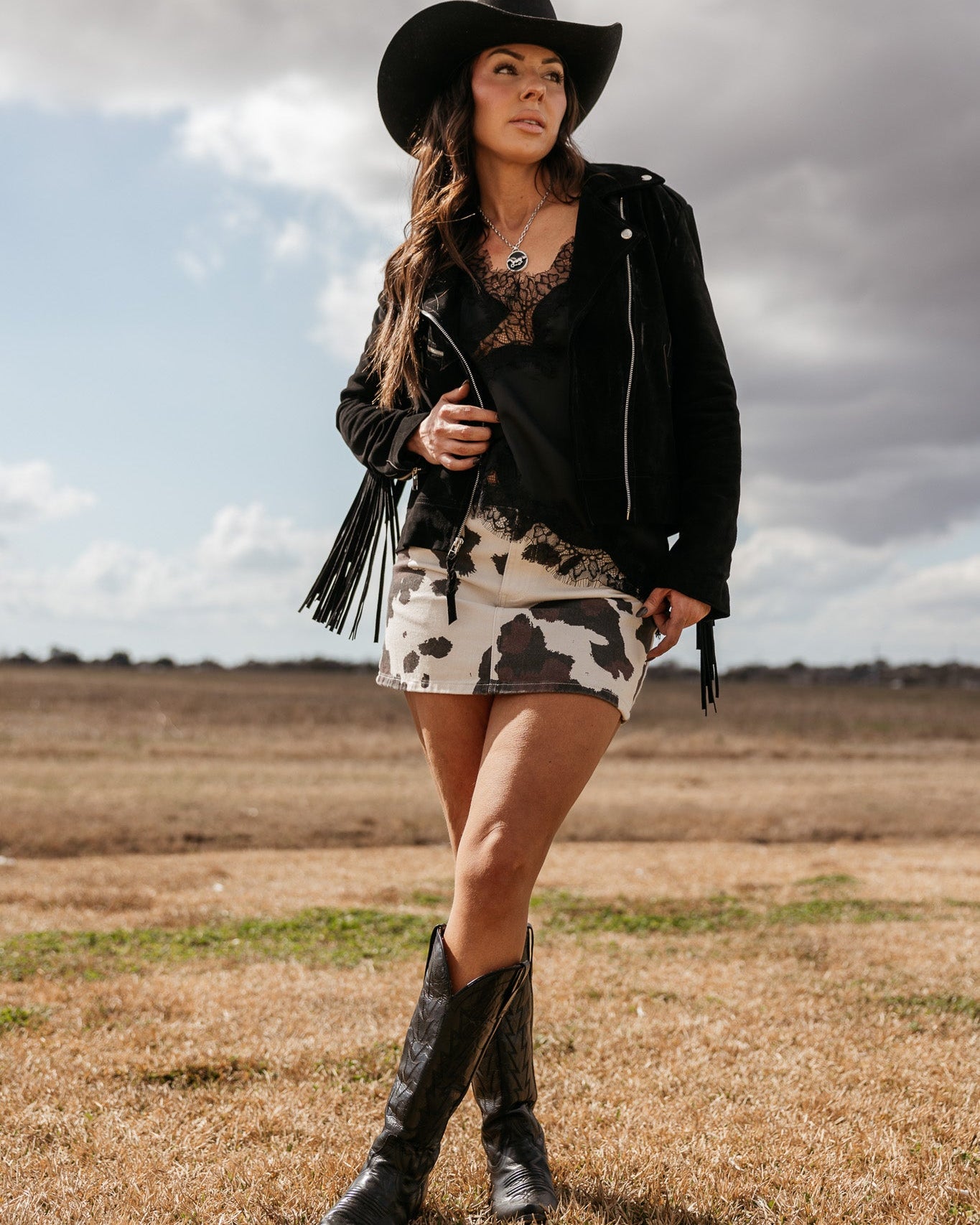 Woman in western cowgirl outfit with black hat, fringed jacket, cow print skirt, and cowboy boots outdoors