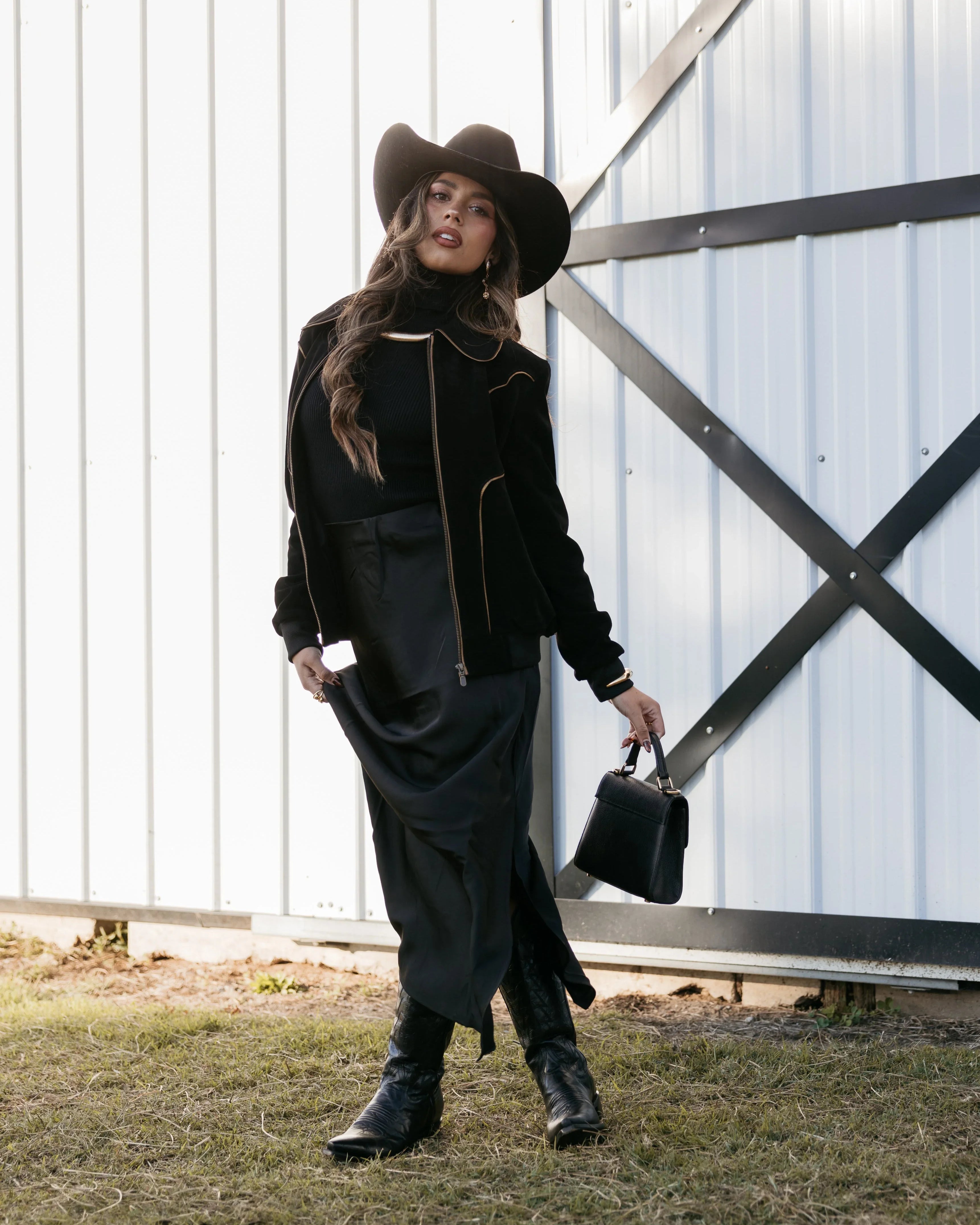 Woman in black cowgirl hat, western jacket, black dress, and boots standing outside barn