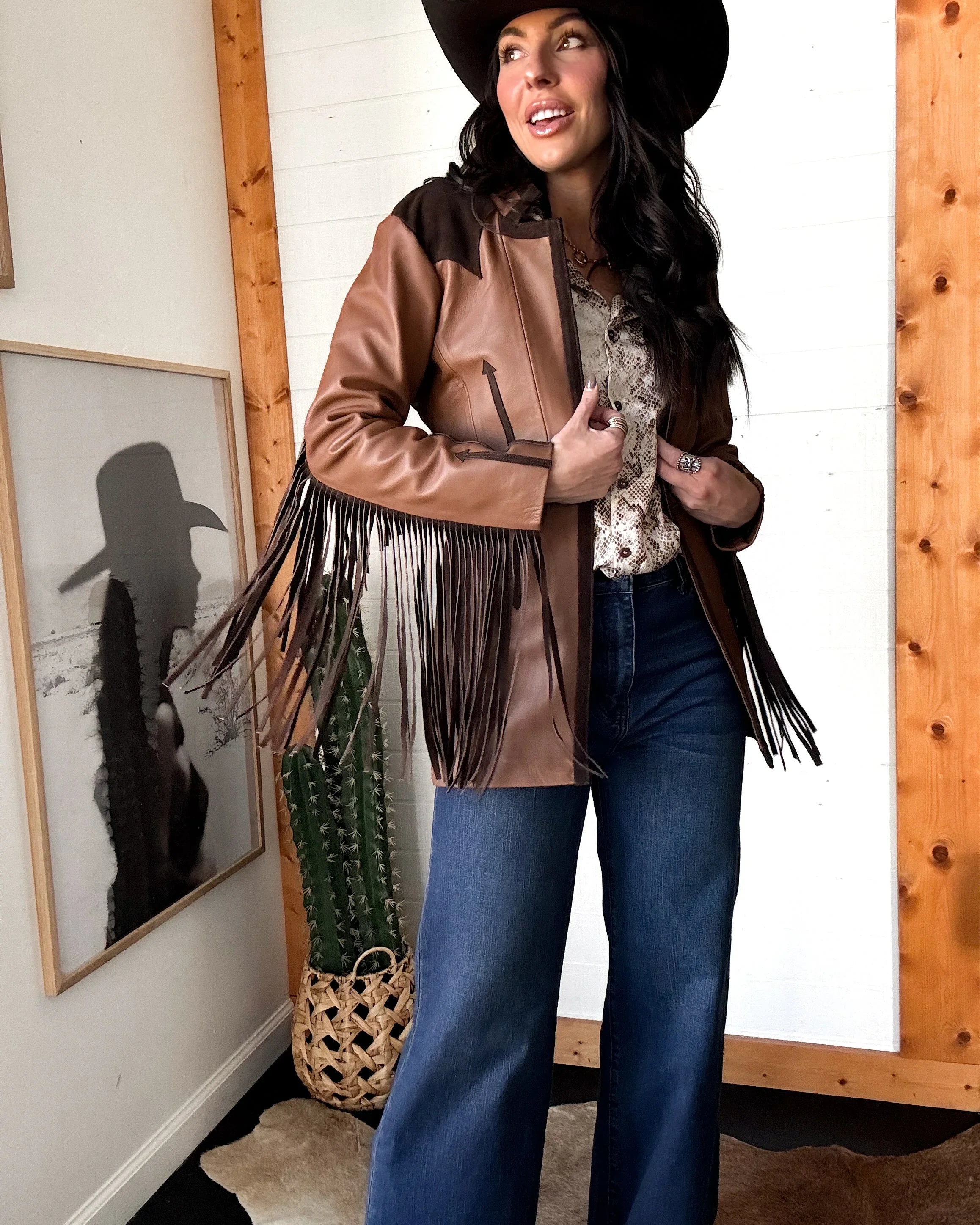 Woman in western fringe leather jacket, wide leg jeans, and black cowboy hat indoors
