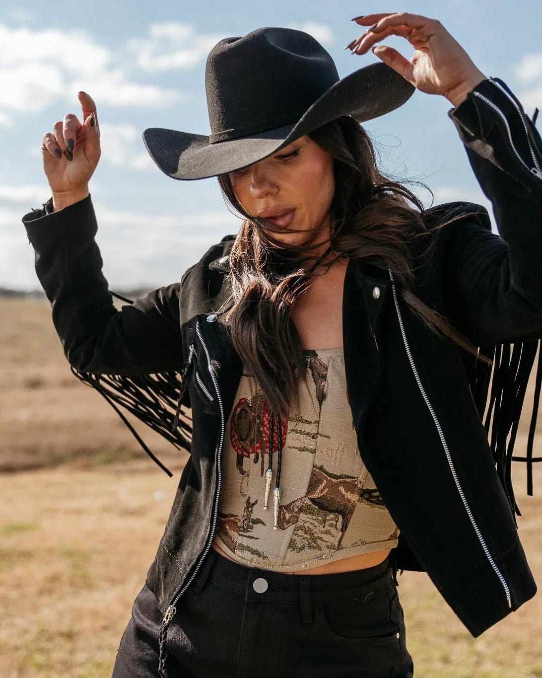 Woman in a black cowboy hat and fringe western jacket outdoors, boho cowgirl outfit style