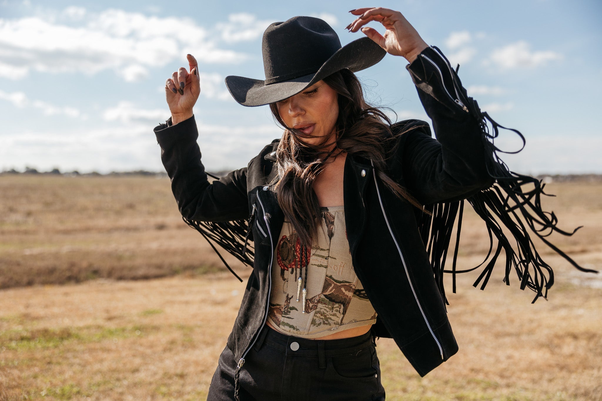 Woman in a black cowboy hat and fringe western jacket outdoors, boho cowgirl outfit style