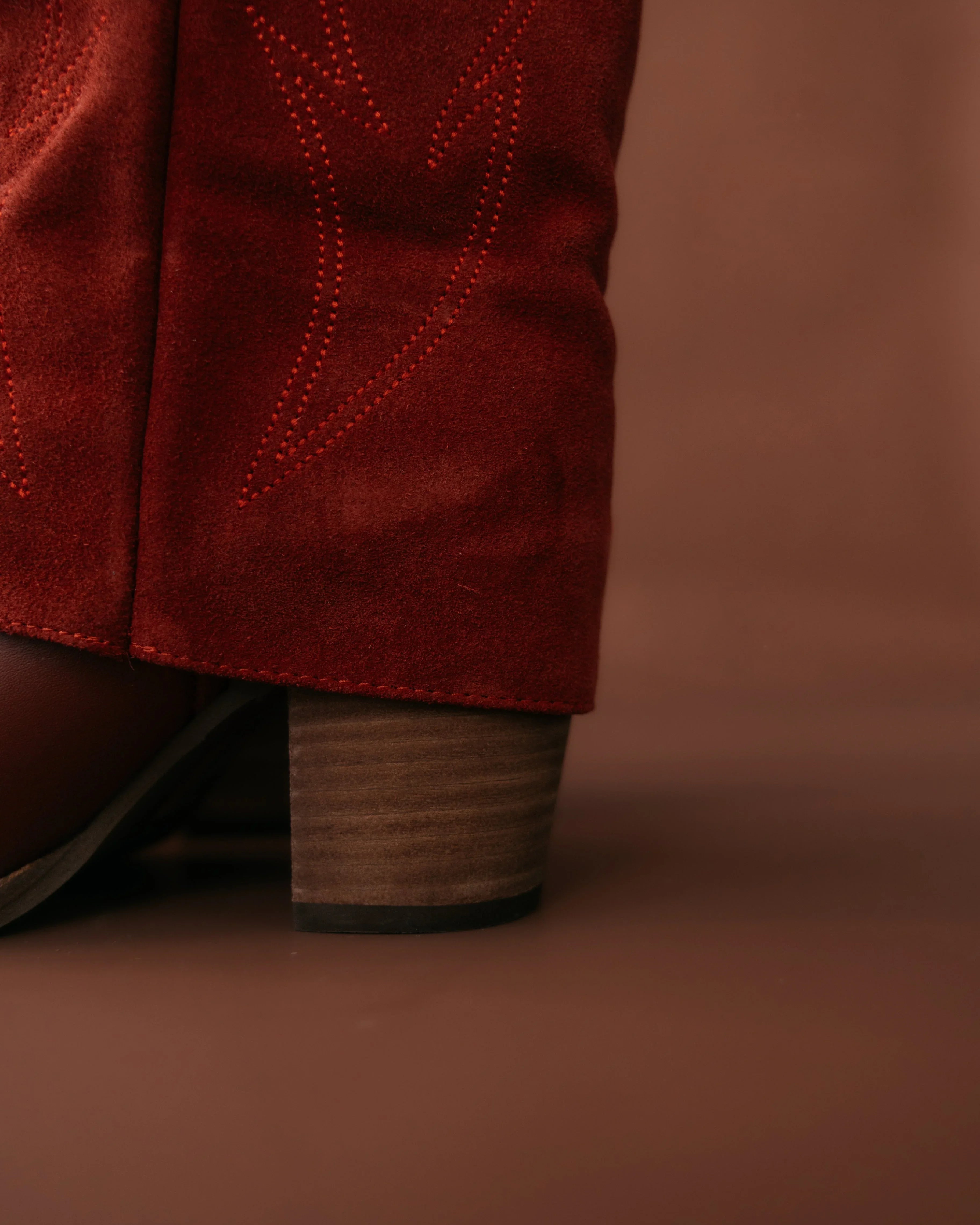 Close-up of red suede western boot with stitched detailing and wooden heel on brown background
