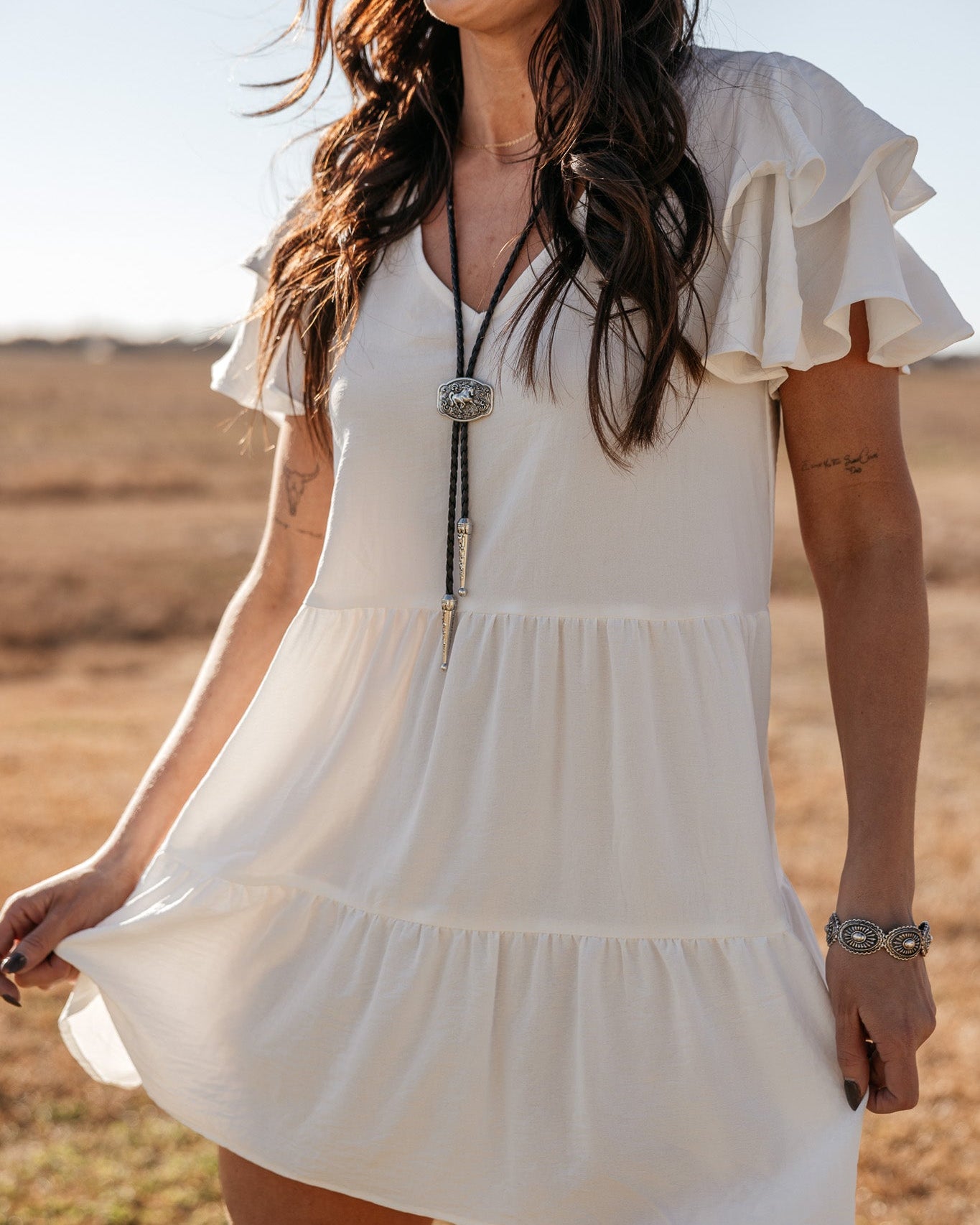 Woman in white ruffle western dress with bolo tie necklace and cowboy hat outdoors