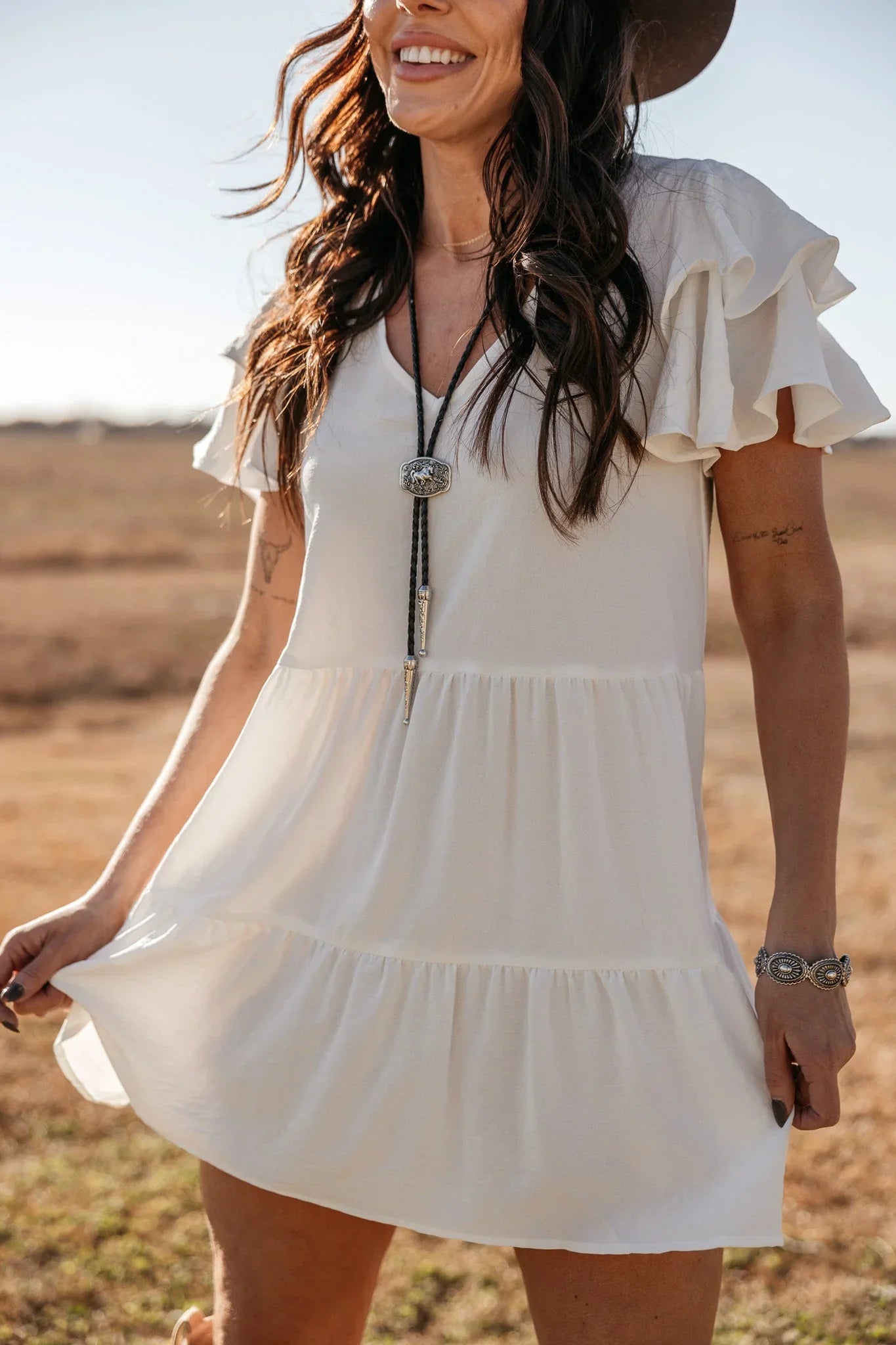 Woman in white ruffle western dress with bolo tie necklace and cowboy hat outdoors