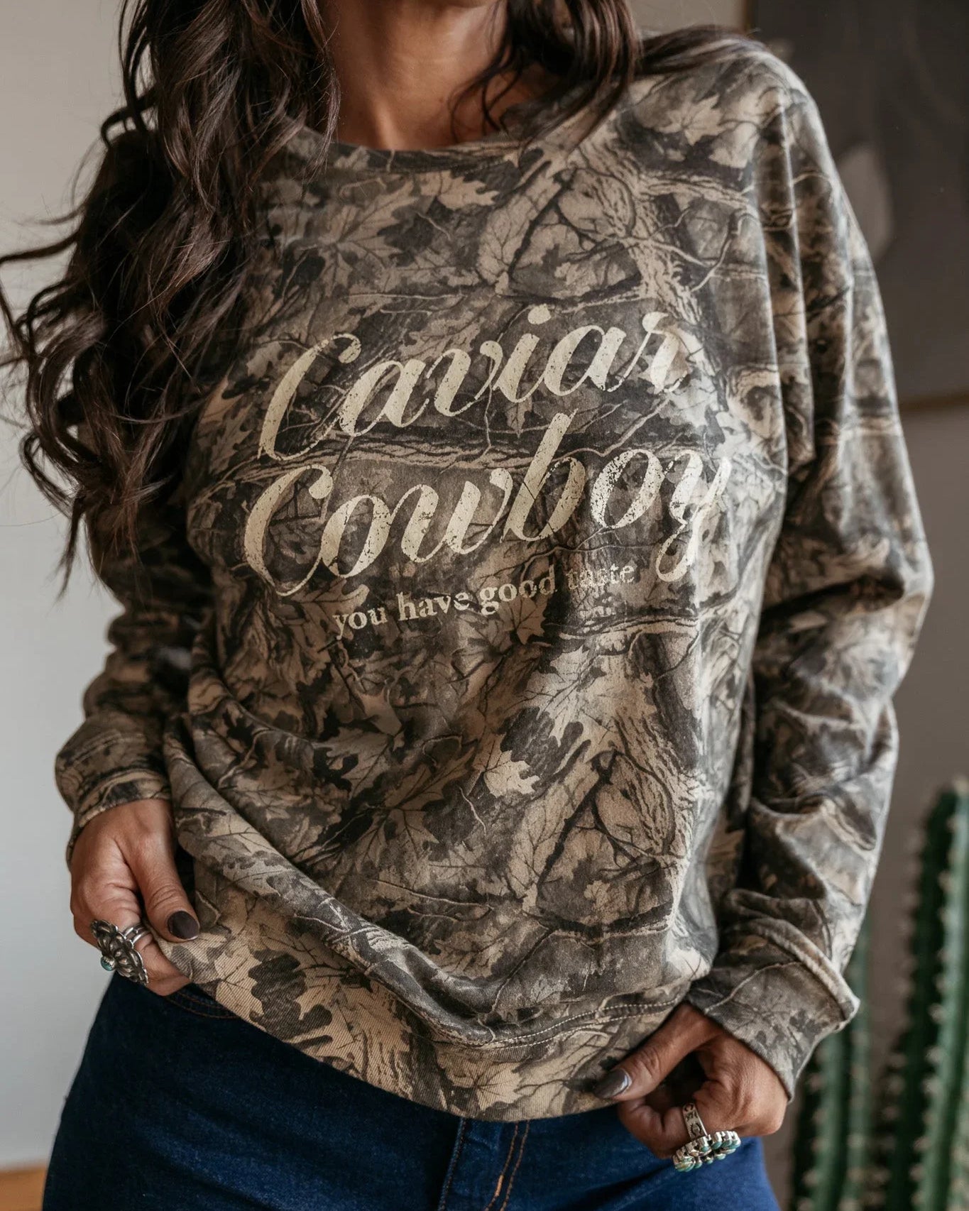 Woman wearing a western boho camo print "Caviar Cowboy" graphic sweatshirt and denim jeans