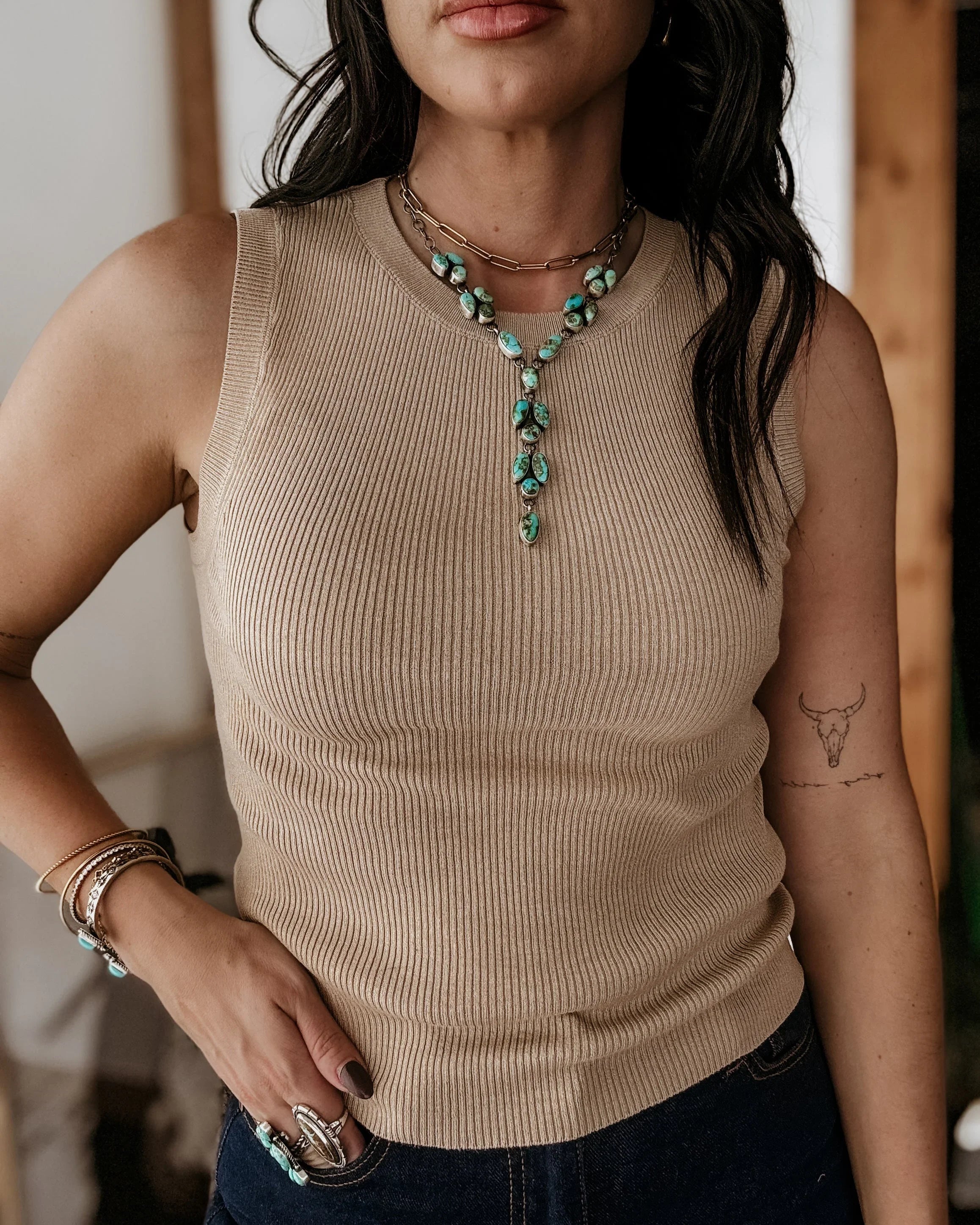 Woman wearing a beige sleeveless top with a green necklace, standing indoors.