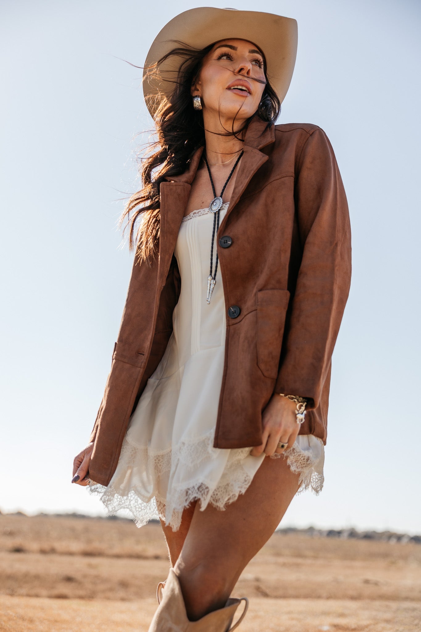 Woman in a western hat, brown suede jacket, white lace dress, and cowboy boots outdoors