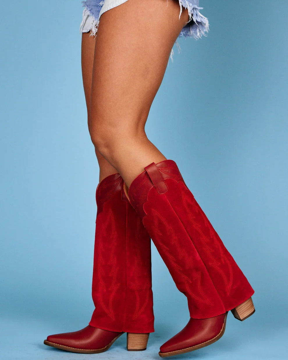 Red western cowgirl boots with wood heel styled with denim shorts on blue background