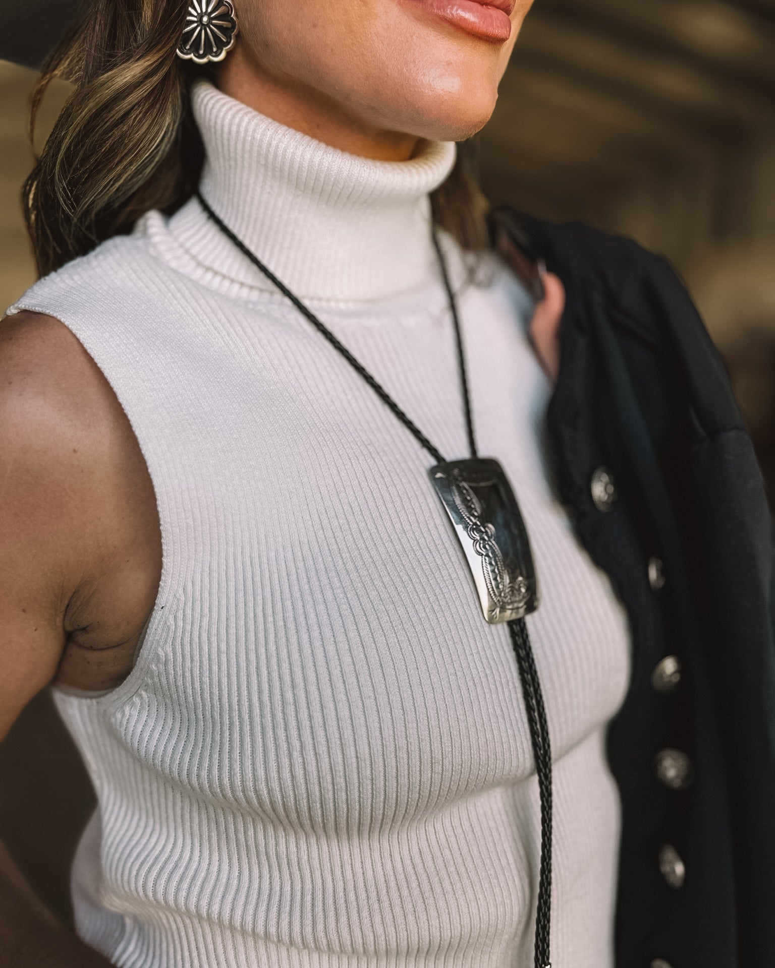 Woman wearing a white ribbed sleeveless turtleneck, western bolo tie, and floral earrings