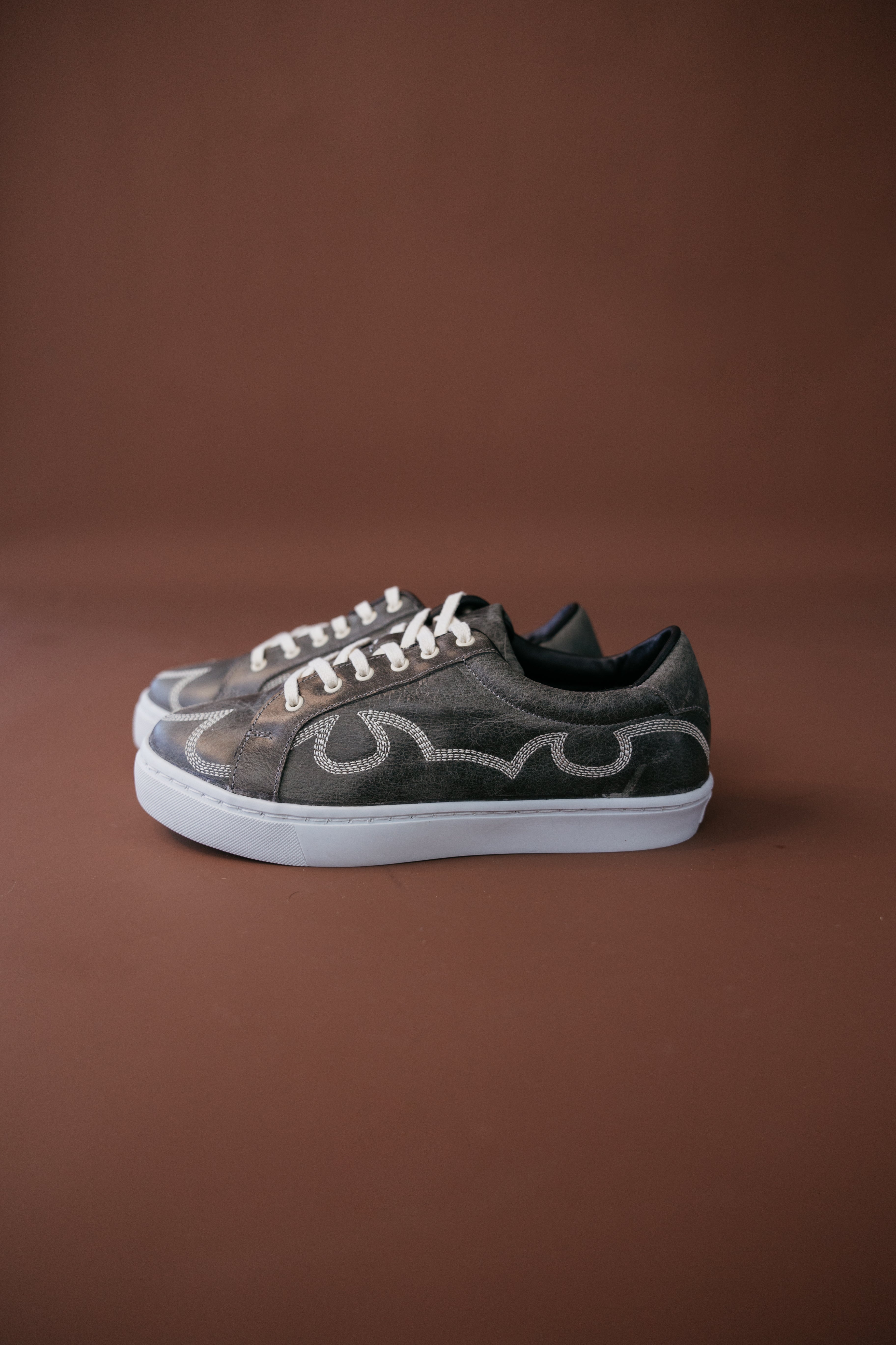 Western-inspired brown sneakers with decorative stitching and white soles on brown background