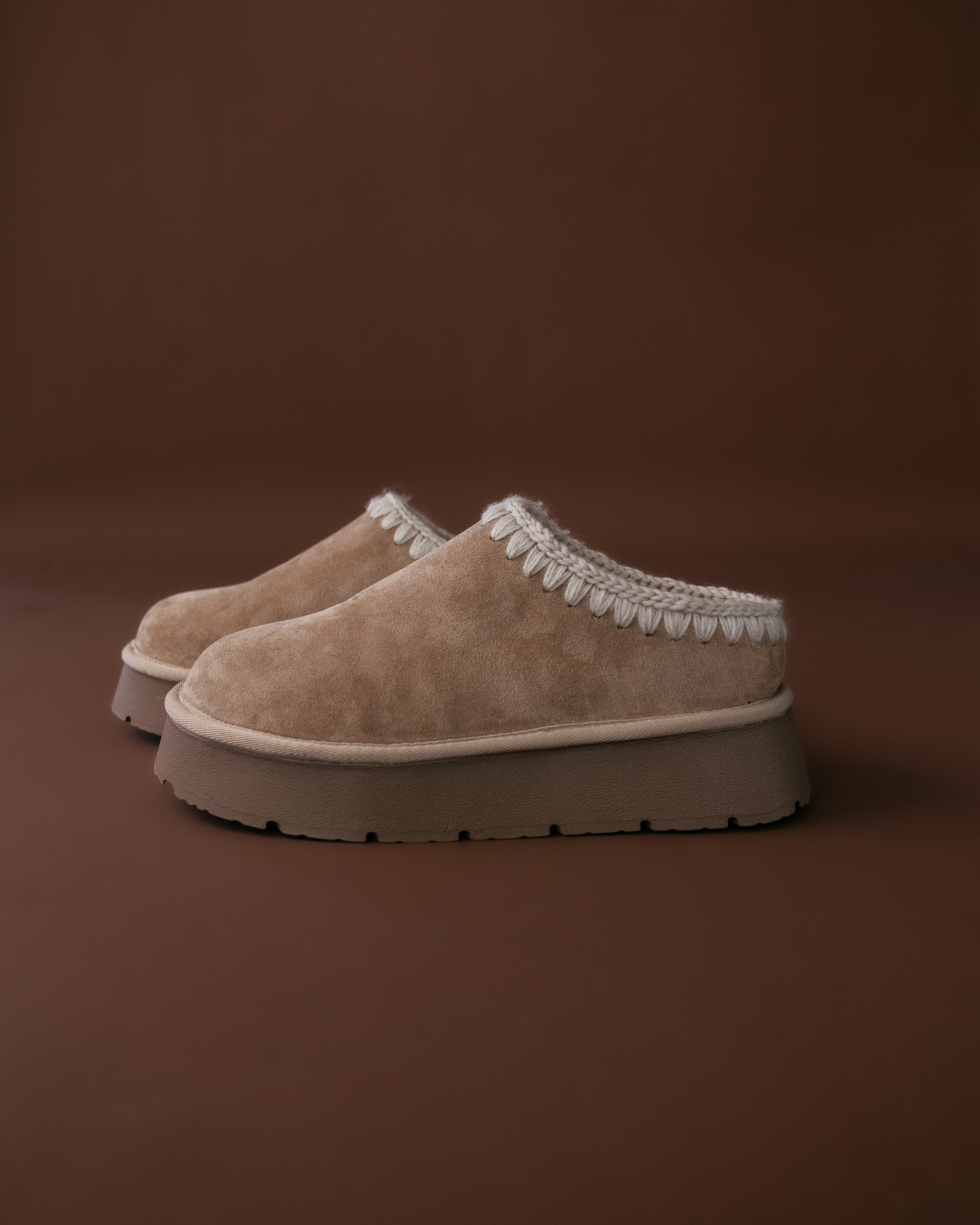 Pair of beige slippers with white trim on a brown background