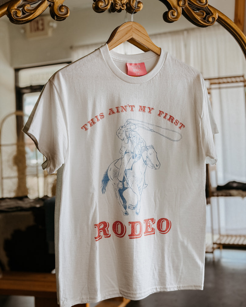 White t-shirt with red and blue graphic and text on a hanger in front of a mirror.