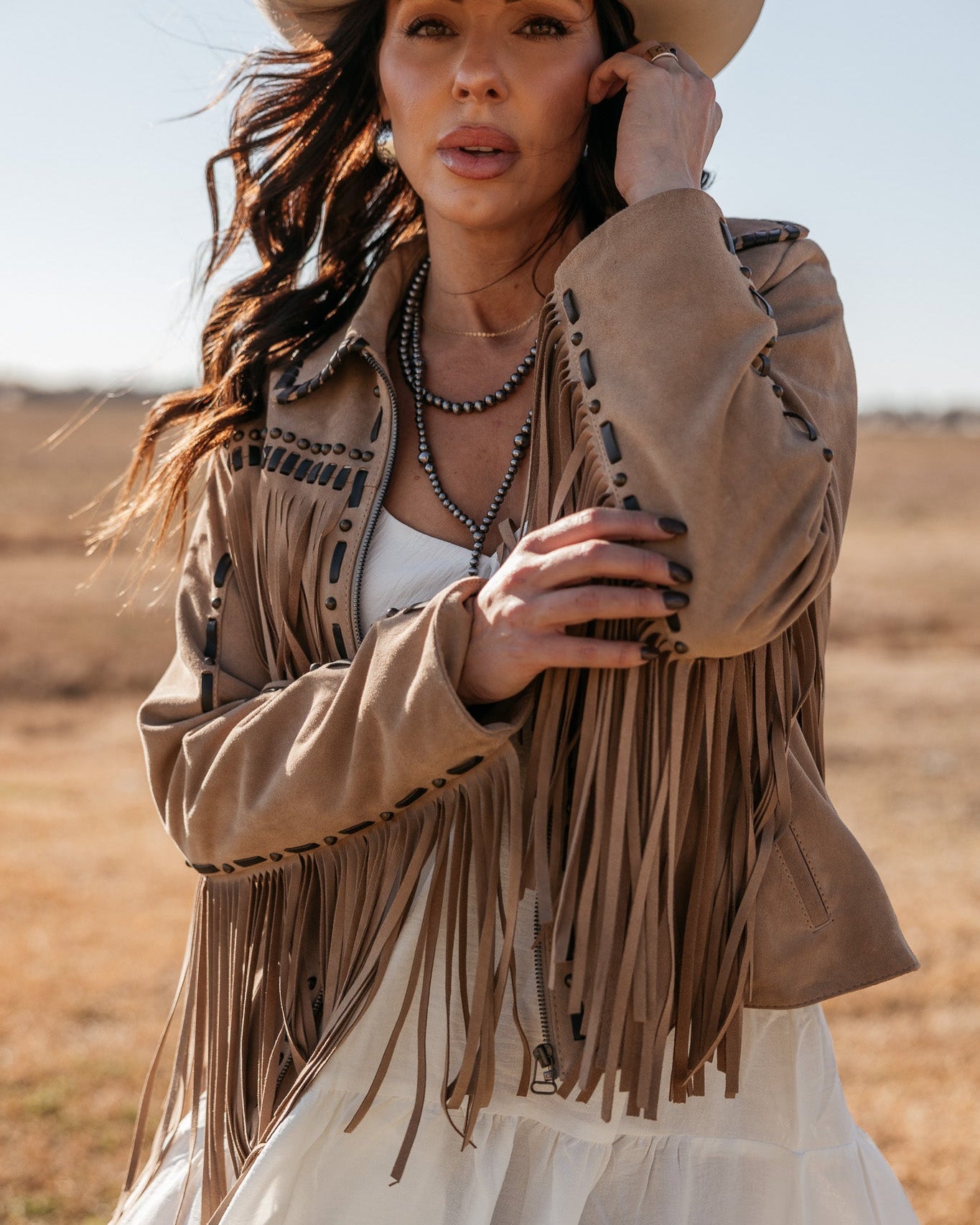 Woman in tan fringe suede jacket, white dress, and cowboy hat in western boho fashion style outdoors
