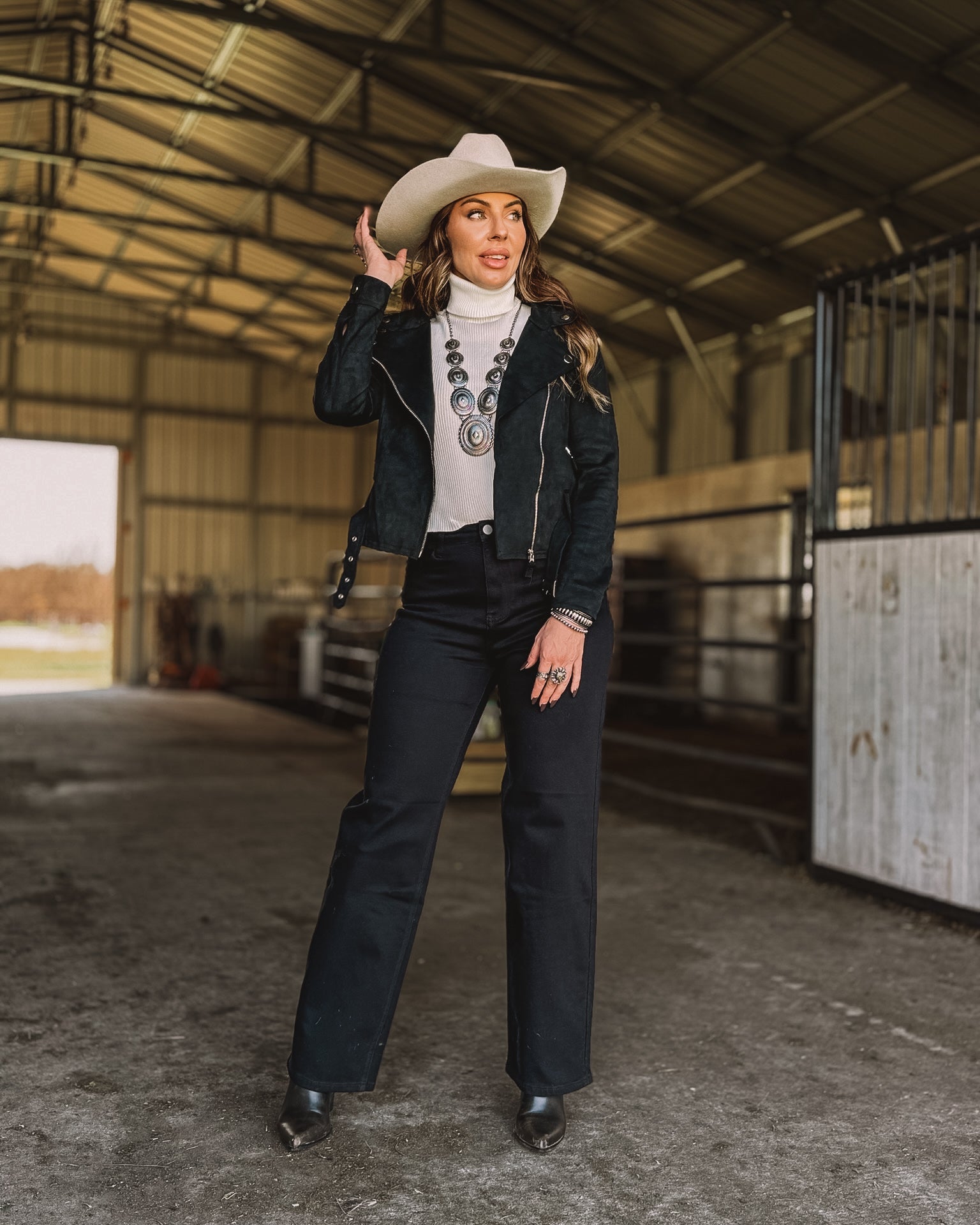 Woman in a white cowboy hat, western jacket, statement turquoise necklace, and black pants in a barn setting