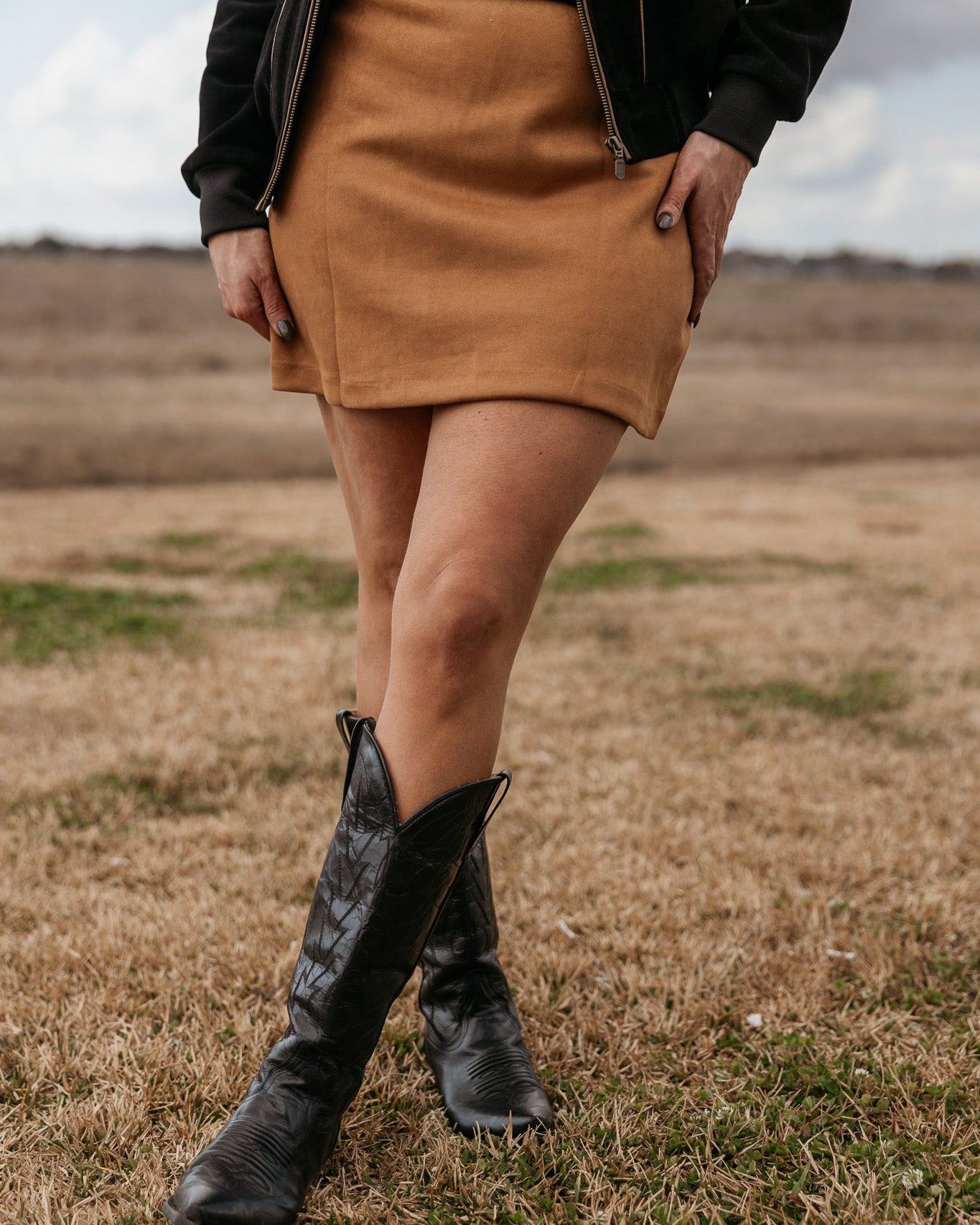 Woman wearing tan mini skirt and black western cowboy boots in a rustic outdoor setting