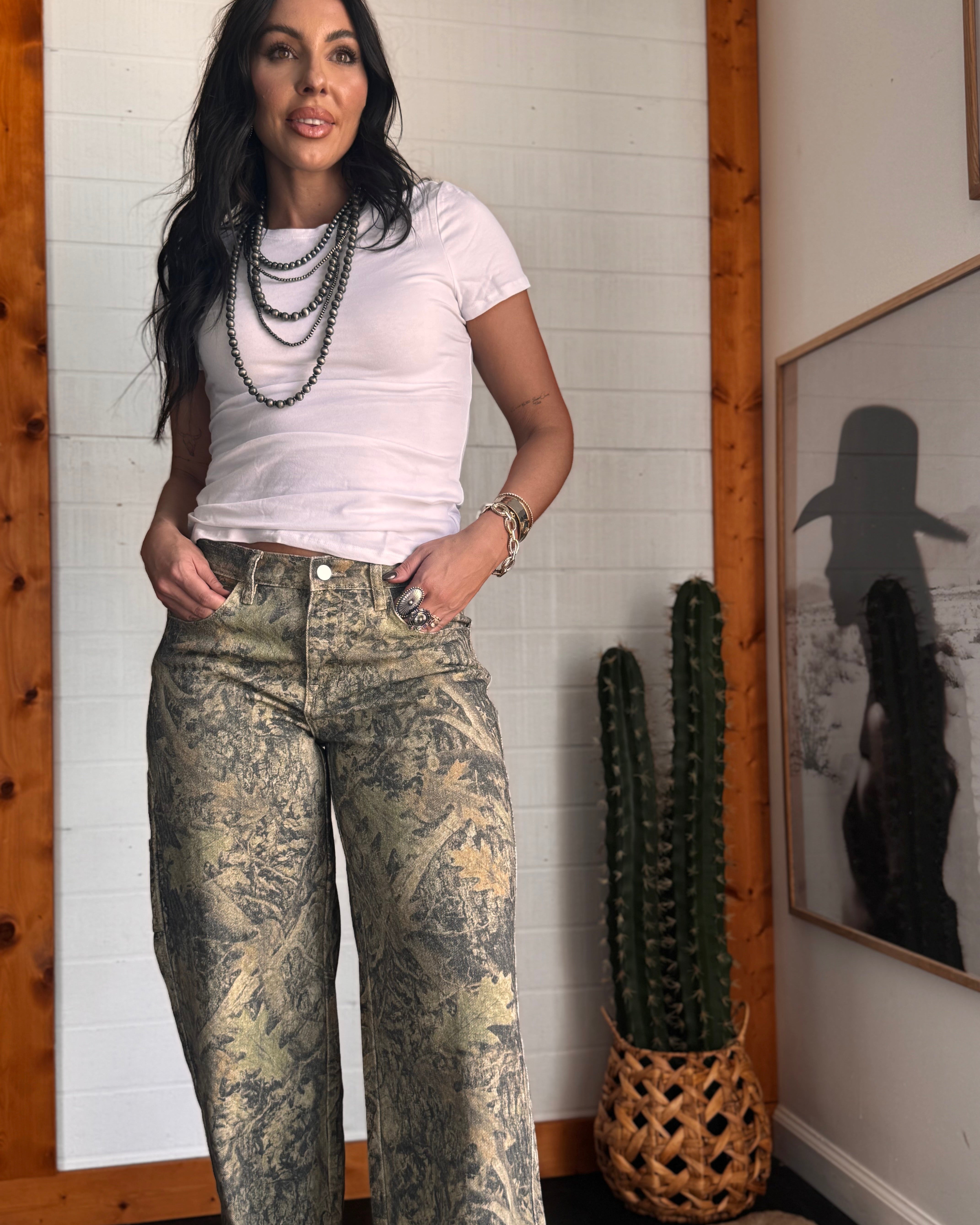 Woman wearing a white t-shirt and patterned jeans standing in a room with a cactus plant and framed picture on the wall.